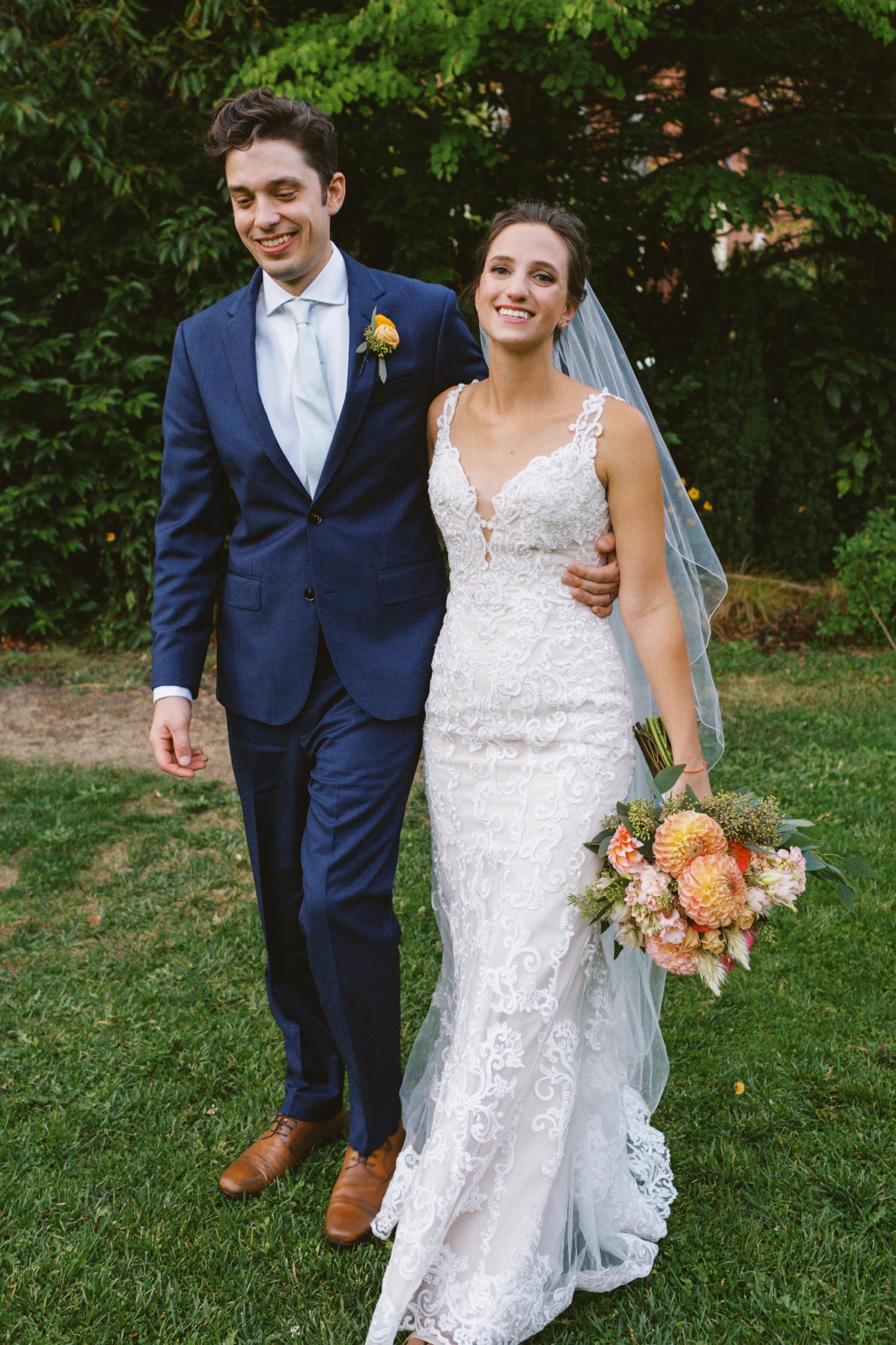 groom in navy blue suit and bride in white dress holding bouquet of pink and orange flowers walk together 