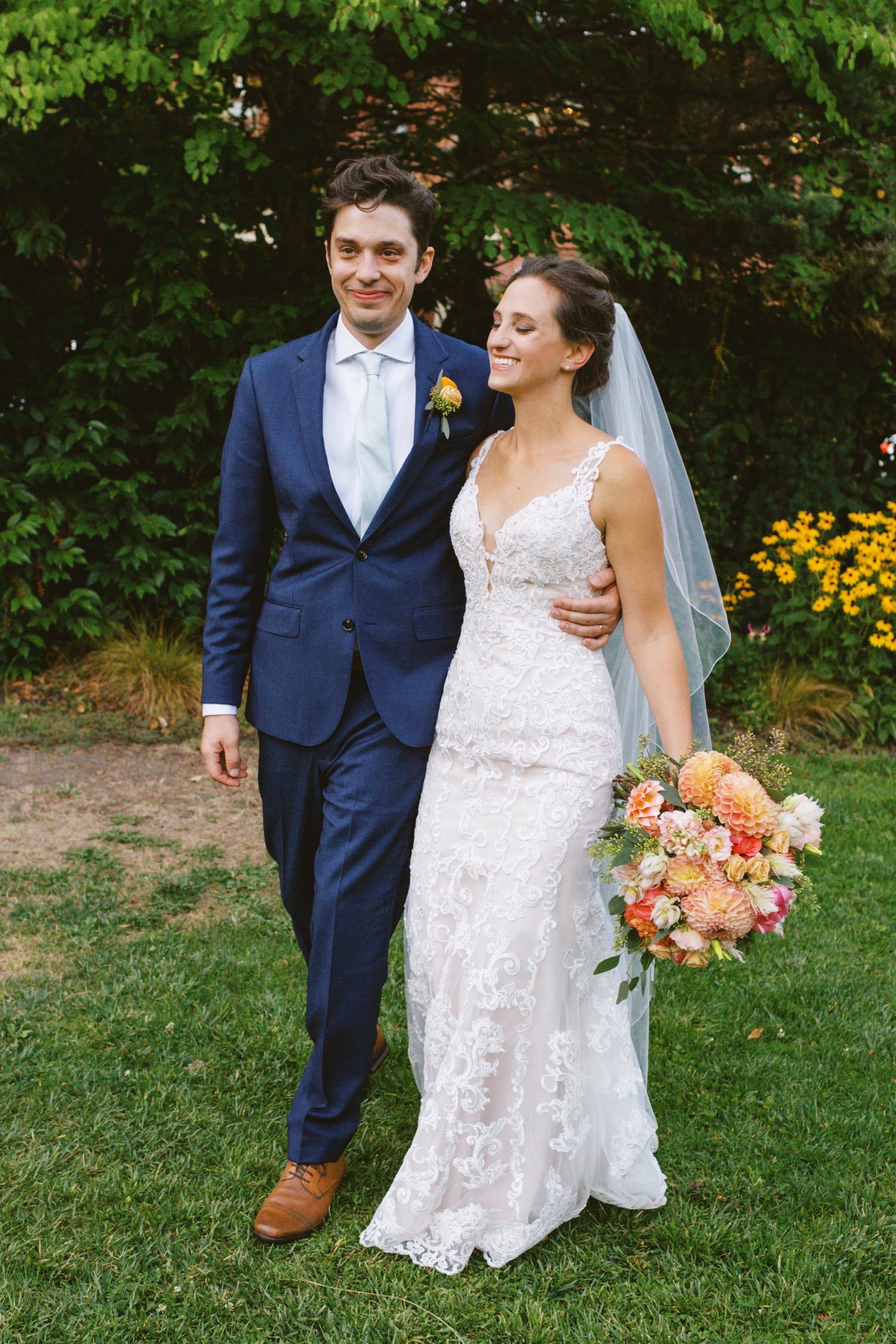  groom in navy blue suit and bride in white dress holding bouquet of pink and orange flowers walk together 