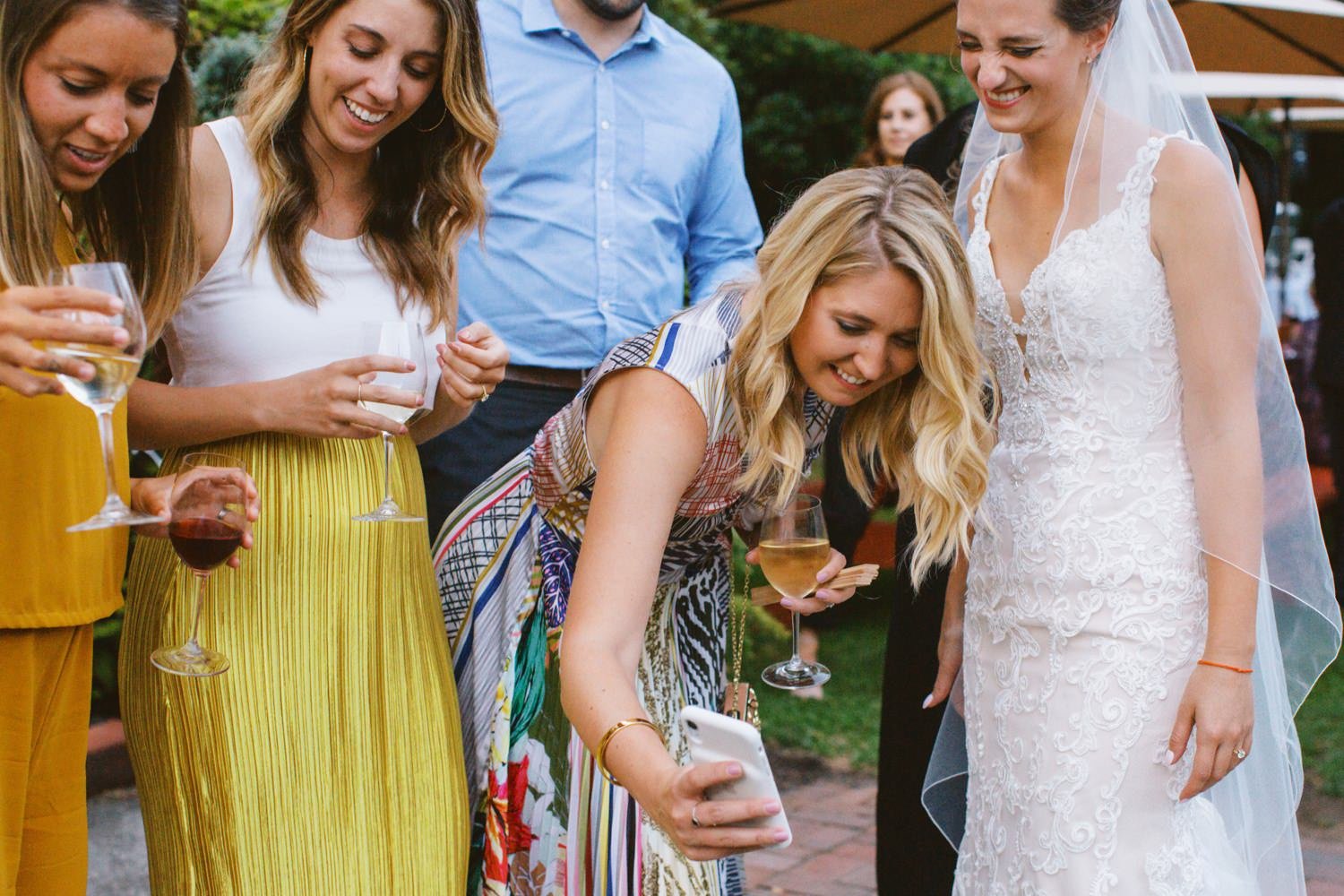  woman holding wine glass and cell phone takes photo while surrounded by women smiling and laughing 