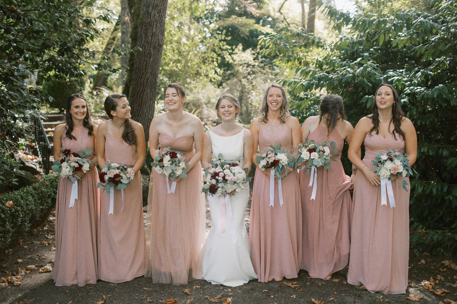  bride in white dress and six bridesmaids in blush pink dresses stand next to each other and laugh while holding bouquets of white and red flowers 