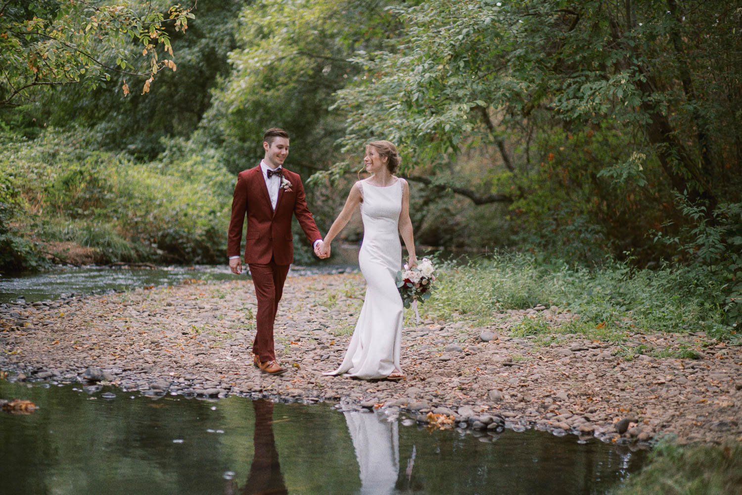  bride in white dress and groom in maroon suit walk hand in hand in front of stream 