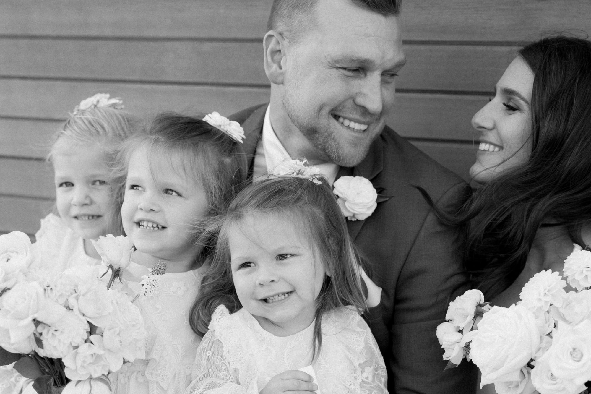  bride and groom look at each other and smile while three flower girls sit on groom’s lap 