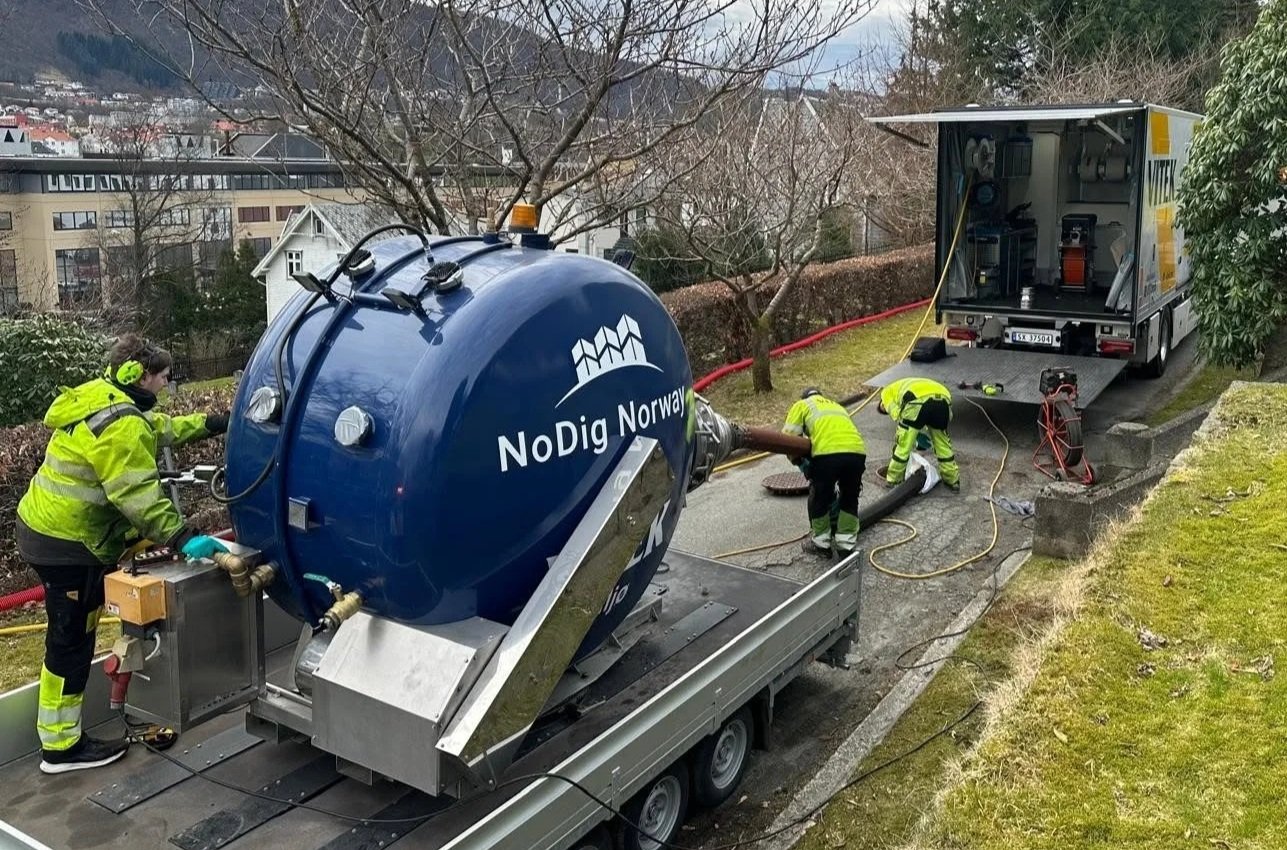 Firemenn installerer en vannpumpe utendørs på en vei, med en stor blå tank merket 'NoDig Norway' og en lastebil i bakgrunnen.