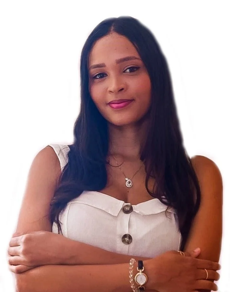 Mulher com cabelo preto e comprido, usando uma blusa branca com botões grandes, acessórios dourados, uma pulseira e um colar, sorrindo com braços cruzados.