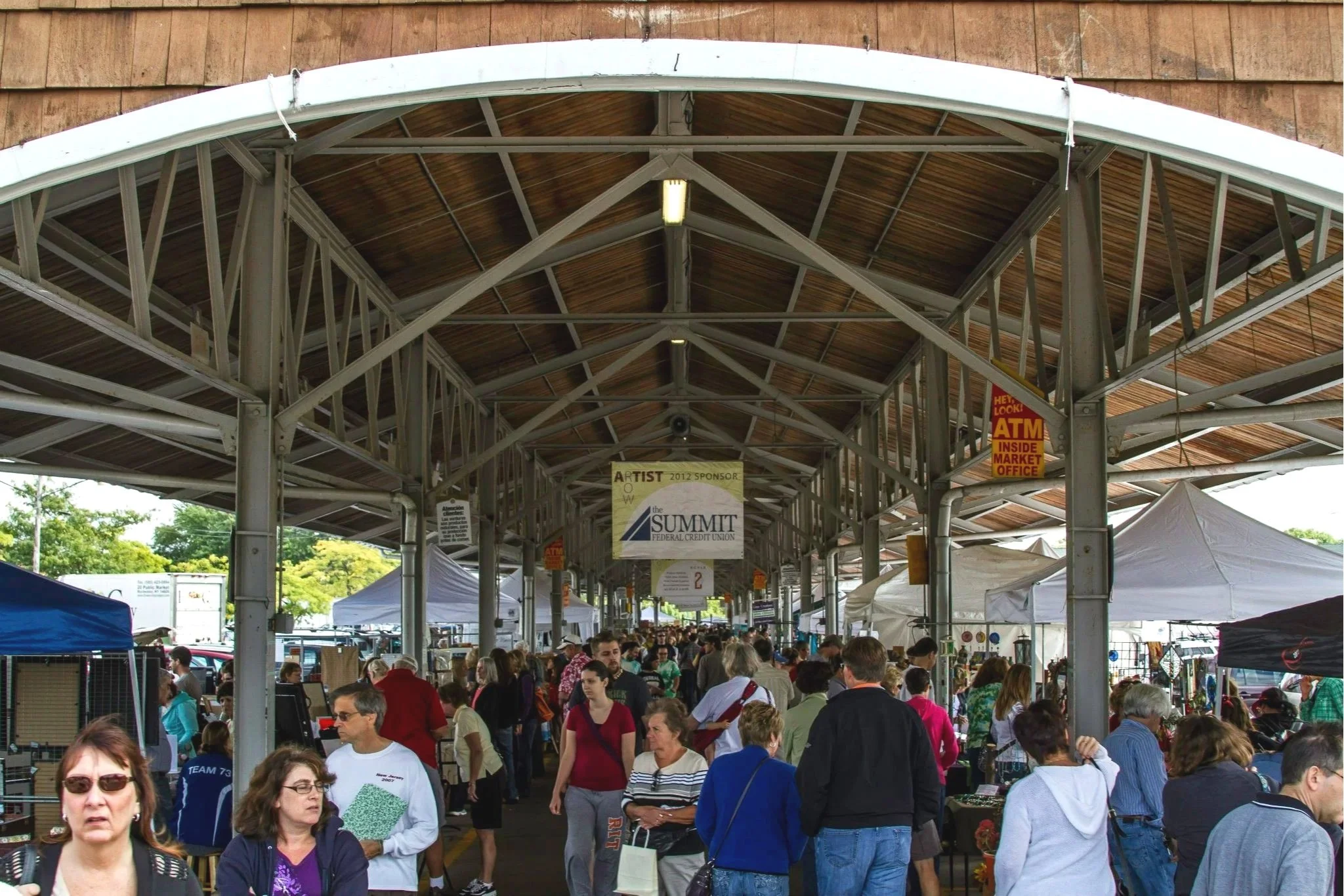 Crowd of people walking and shopping inside a covered outdoor market with vendor tents and signs, including a banner for the Summit Federal Credit Union.