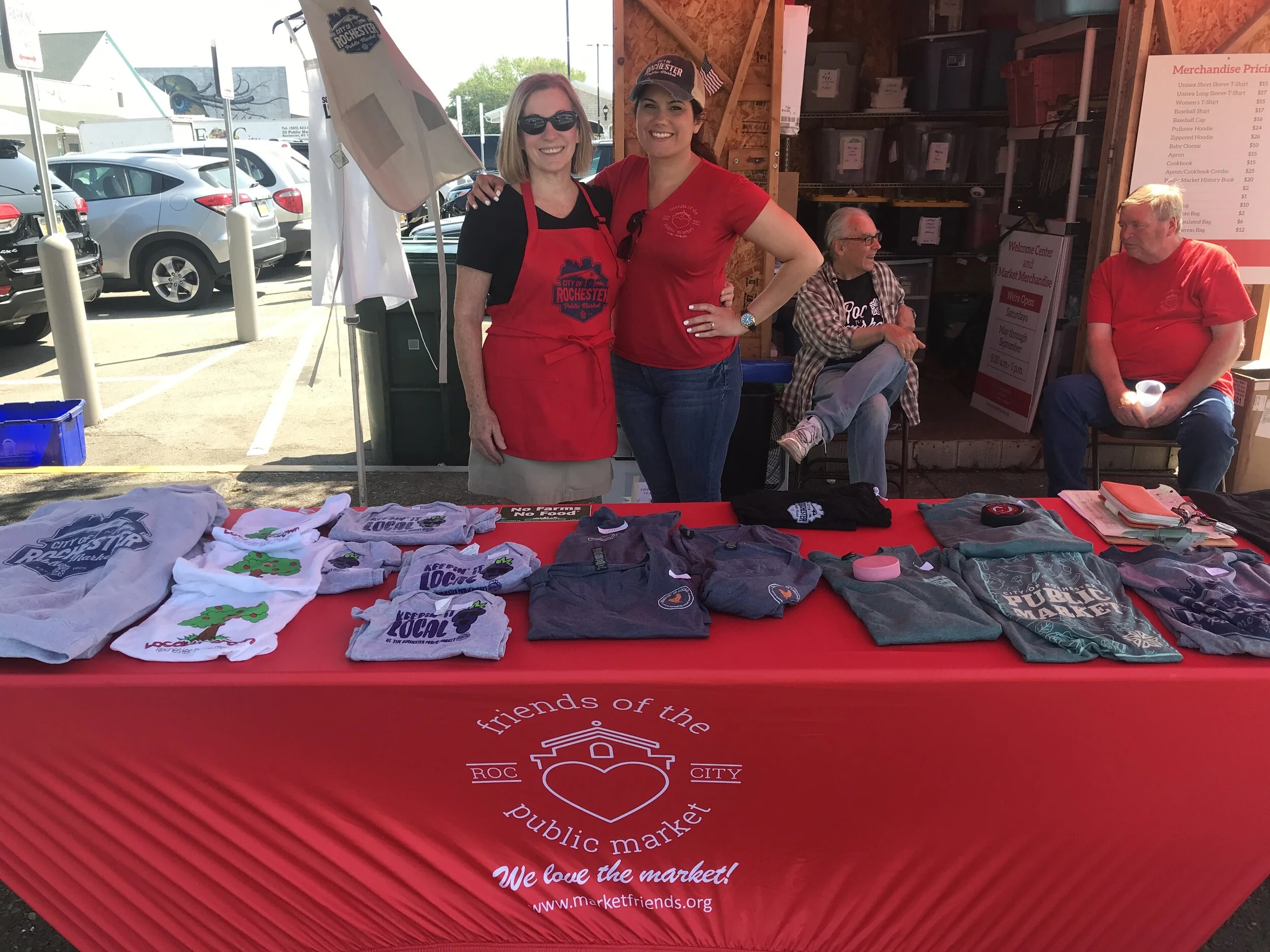 Two women wearing red aprons and shirts, smiling at the camera, standing behind a table at a market booth. The table displays various clothing items like t-shirts, sweatshirts, and hats. In the background, two men are sitting, one with a plaid shirt, and the other with a red shirt. The booth has a red tablecloth with a logo and text for 'Friends of the Public Market' in Rochester, NY.