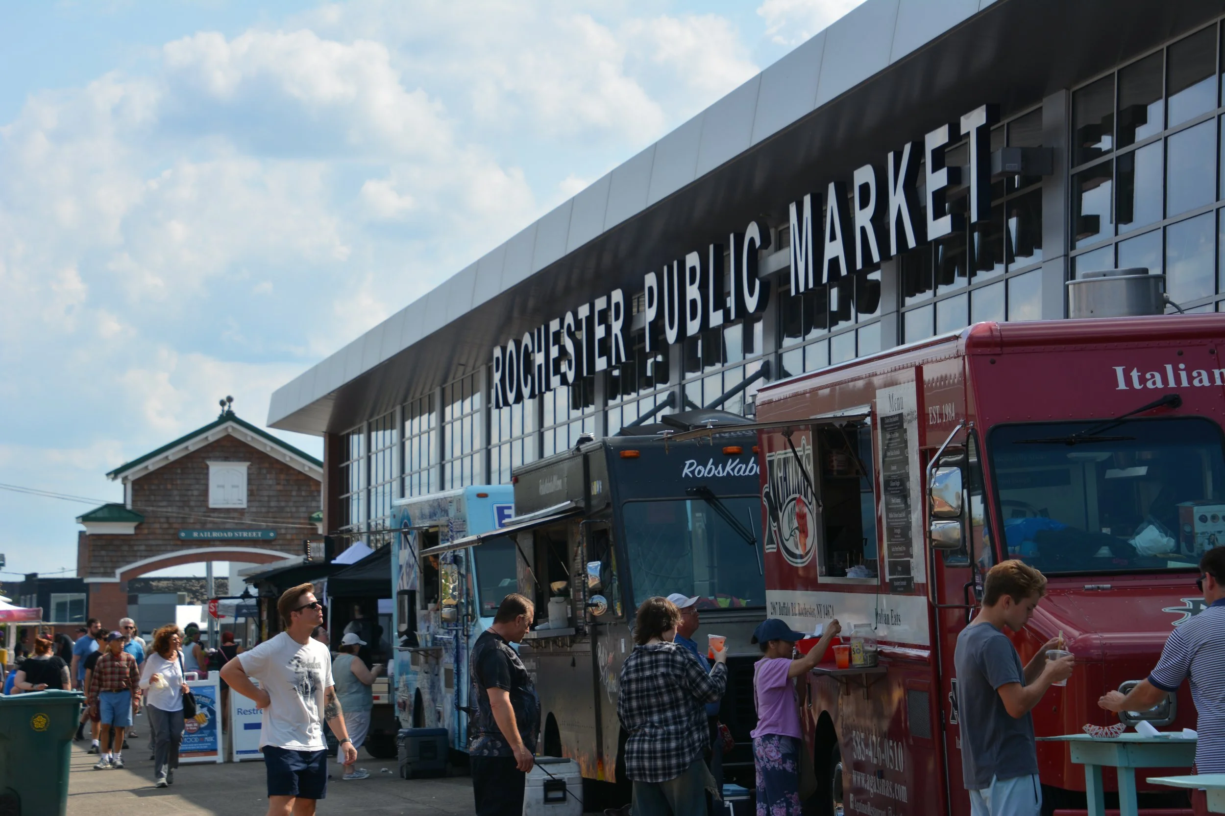 People at food trucks outside Rochester Public Market during daytime with a building and cloudy sky in the background.