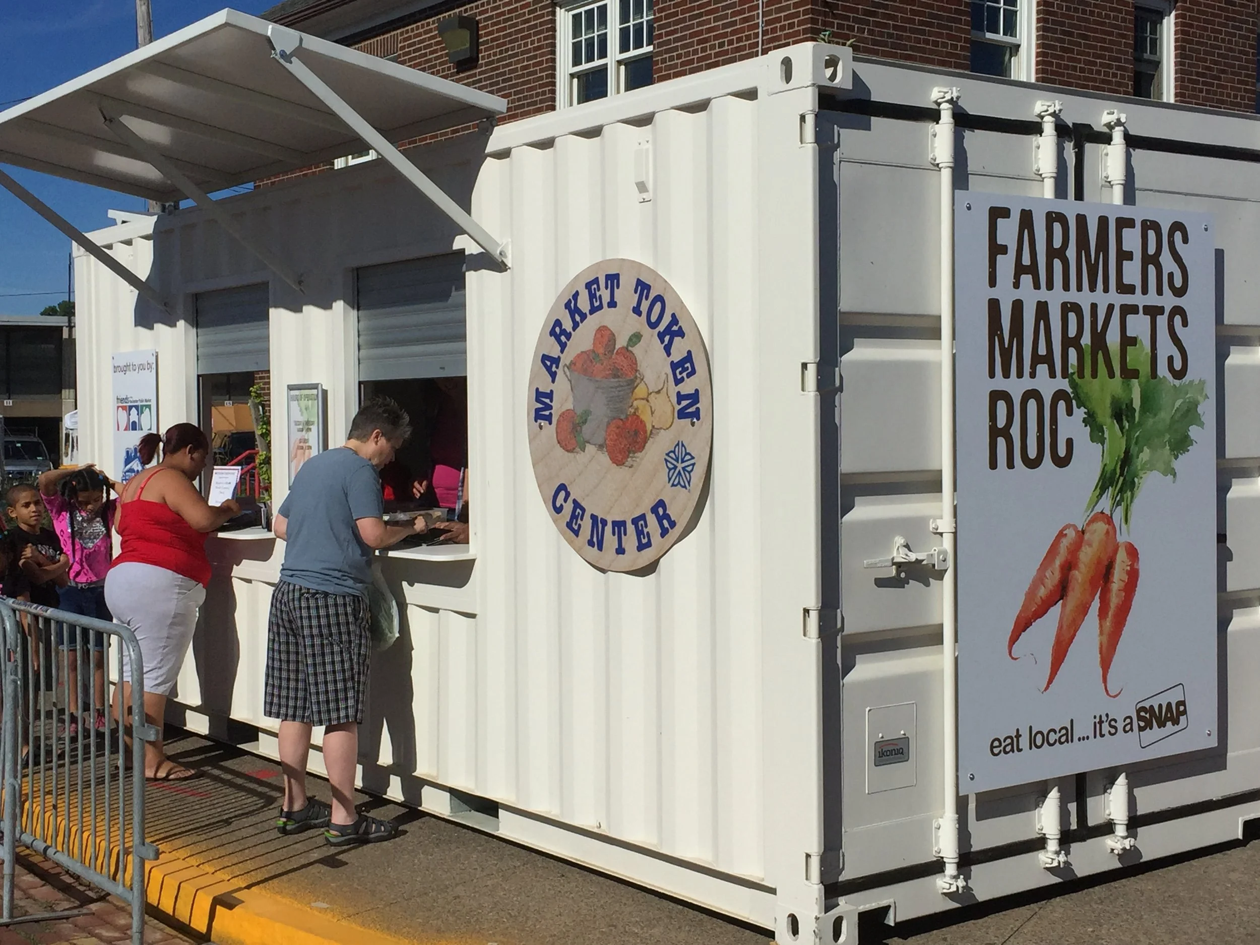 People standing in line at a mobile farmers market stand, which is a white shipping container with signs for 'Farmers Markets Roc' and 'Market Token Center,' and a large sign with carrots and the phrase 'eat local...it's a SNAP.'