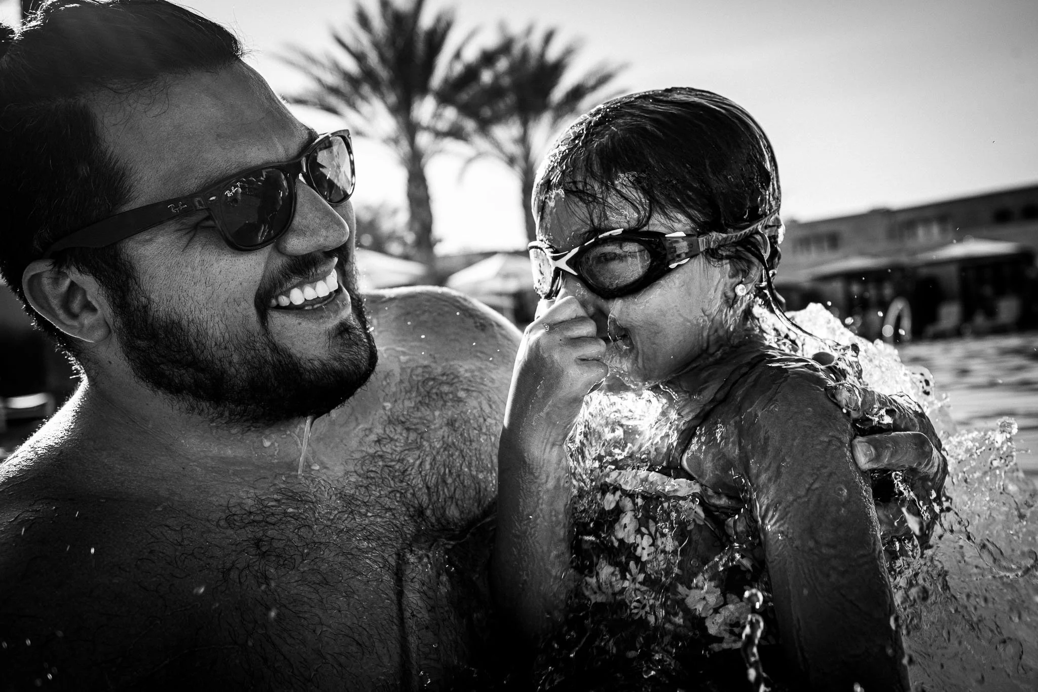 A man and a girl are playing in the pool, both wearing swimming goggles. They are laughing, and the girl is touching her nose. In the background, palm trees and buildings are visible.