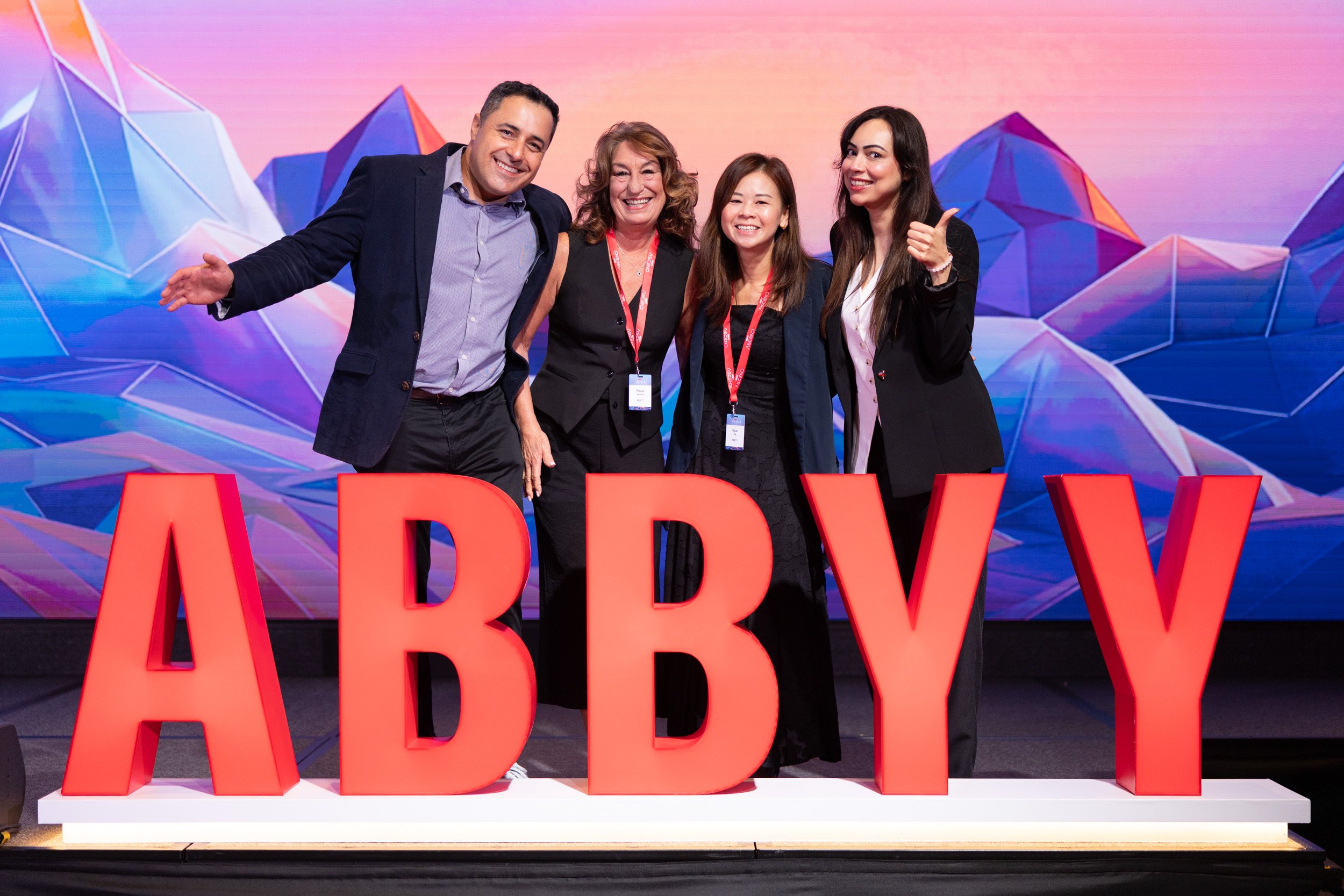  Group portrait of ABBYY staff team posed on stage beside the official company logo at PARKROYAL COLLECTION Pickering. 