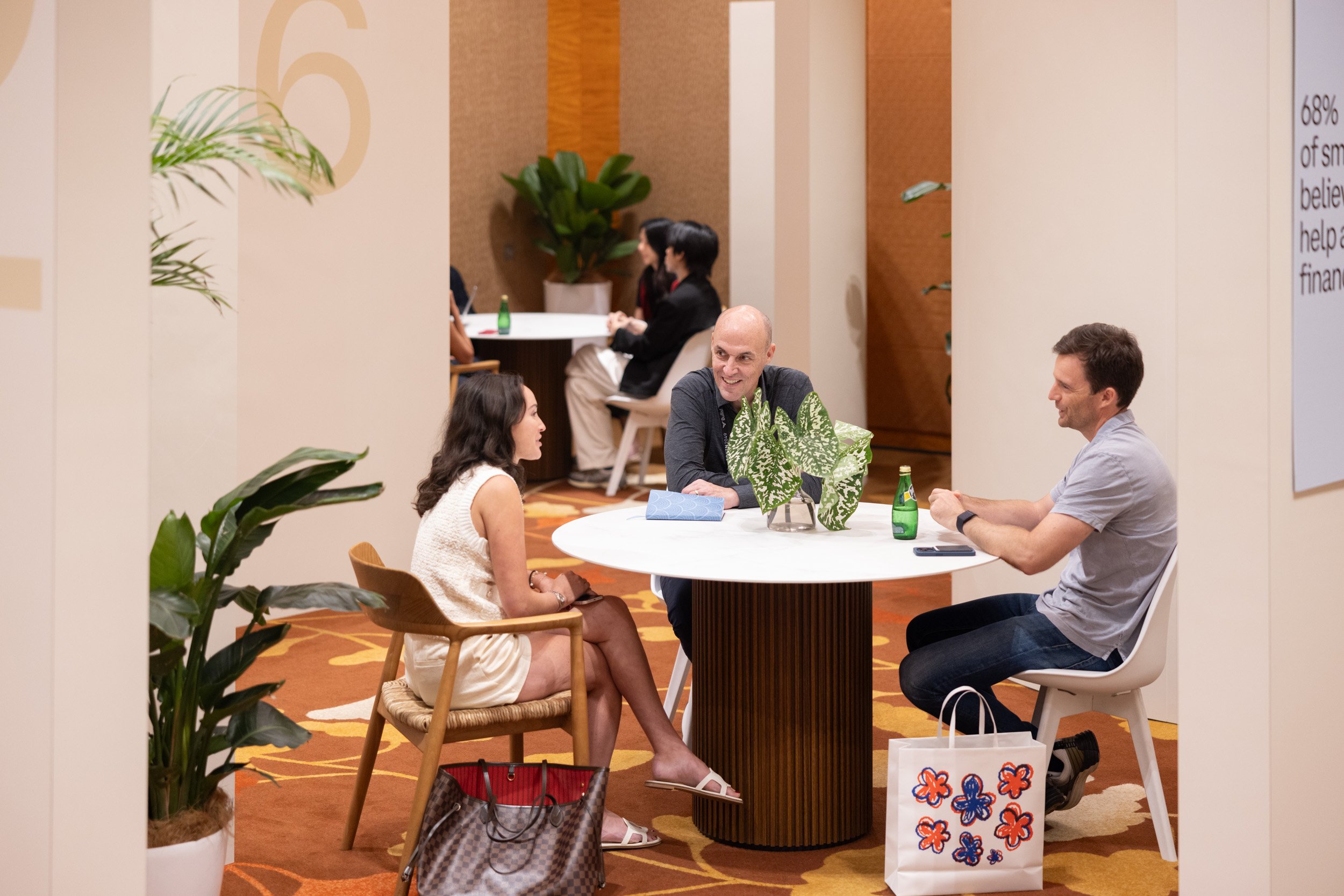  Medium perspective of three delegates engaging in B2B discussions around a meeting table inside numbered exhibition booths. 