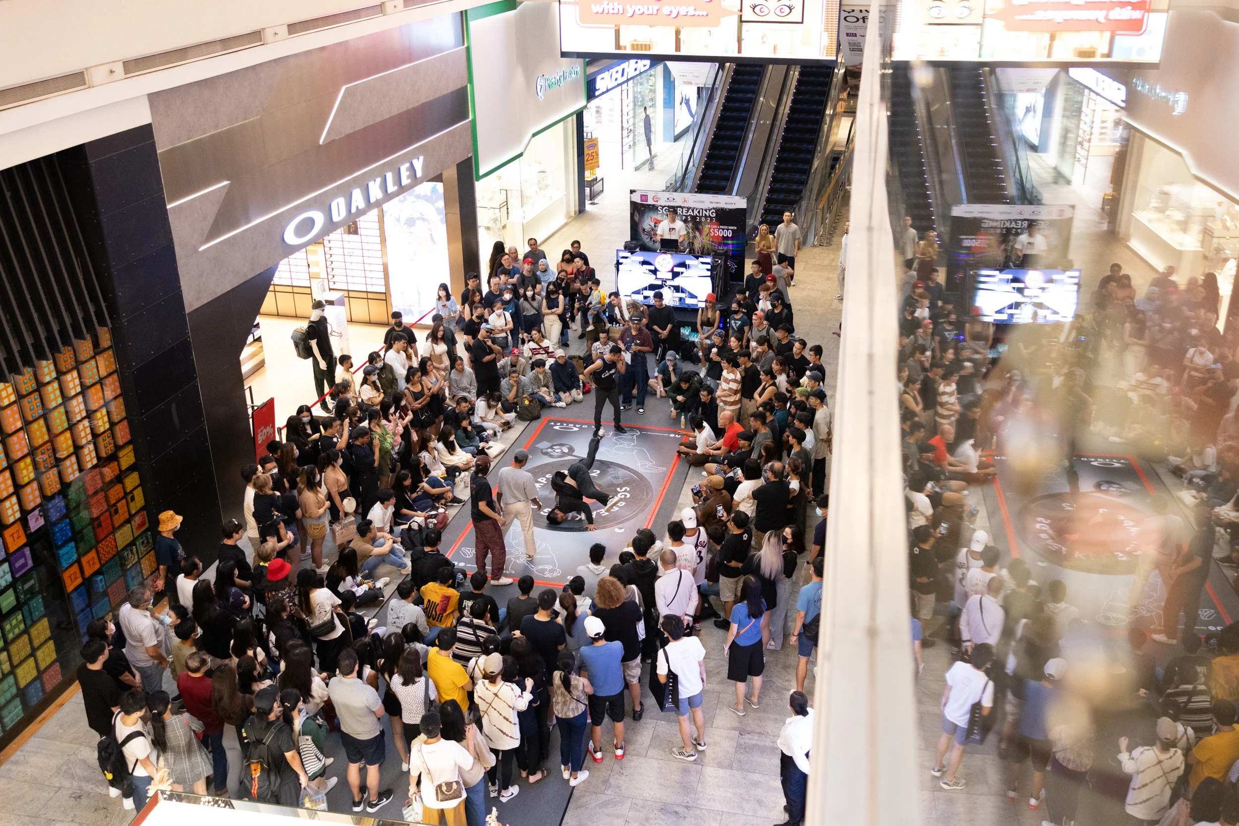  High-angle shot from the second floor showing a B-boy executing a freeze pose on the branded competition mat near the Oakley store at 313@somerset. 