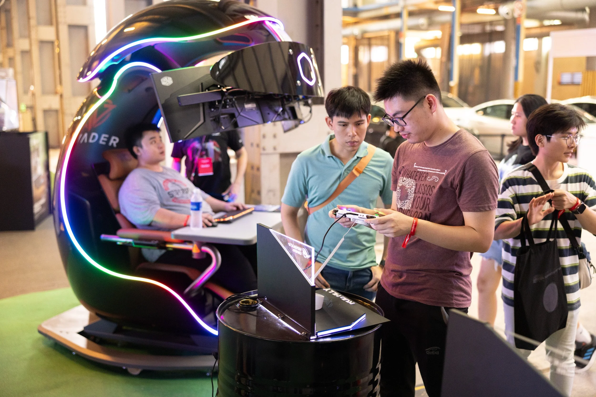  Visitors testing the ASUS ROG Ally handheld console with the Invader PC simulator rig operating in the background. 