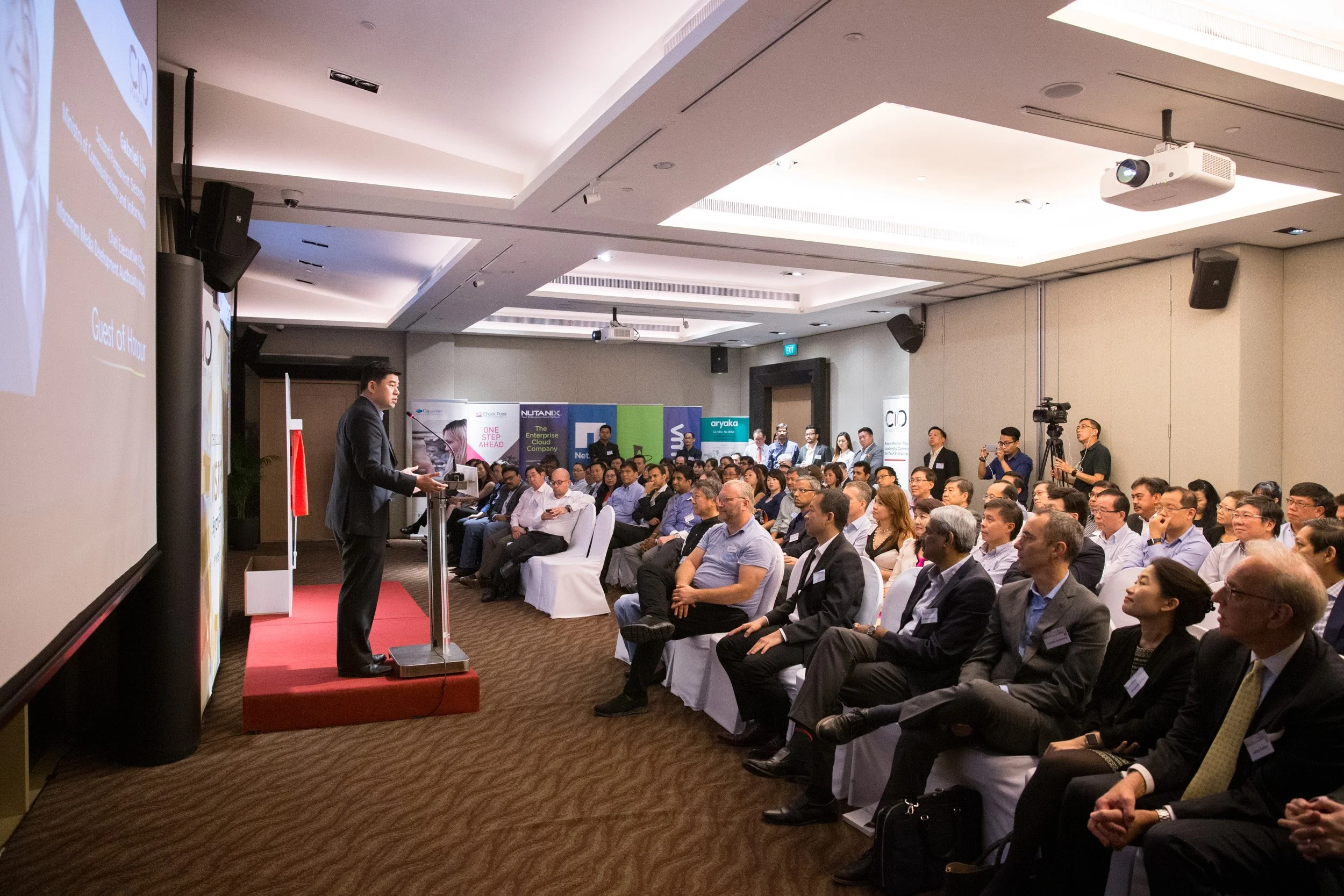  Side-angle view capturing Guest of Honor Gabriel Lim speaking on stage with attentive C-suite audience members in view. 