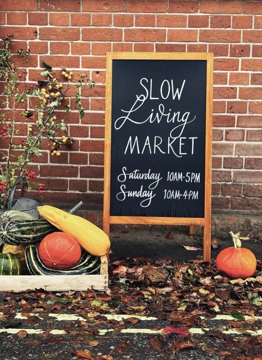 A chalkboard sign outside a brick wall reads "Slow Living Market" with hours listed for Saturday 10AM-5PM and Sunday 10AM-4PM. In front of the sign, there are pumpkins, gourds, and fallen leaves, reflecting an autumn or fall setting.