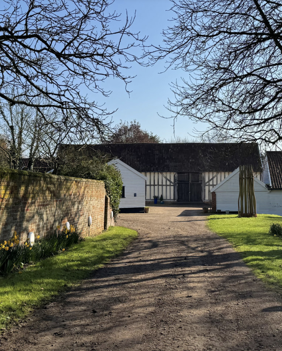 A rural scene with a dirt driveway leading to a large barn with black and white timber framing, flanked by small white sheds on either side. There are grass lawns with daffodils along the brick wall on the left and leafless trees overhead, casting shadows on the ground while a clear blue sky is visible in the background.