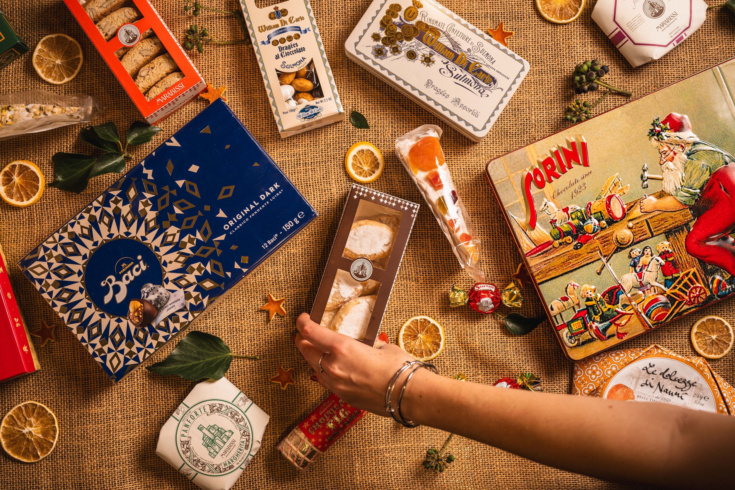 Assorted holiday sweets and cookies on a woven tablecloth, including chocolate boxes, citrus slices, candy, and festive decorations.