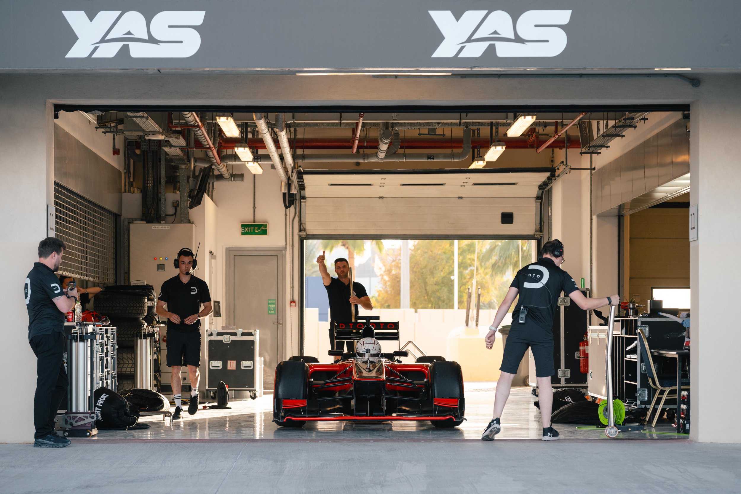 A Formula racing car inside a garage with team members preparing the car, one team member giving a thumbs up, surrounded by equipment and tires, with a partially open garage door revealing a sunny outdoor setting.