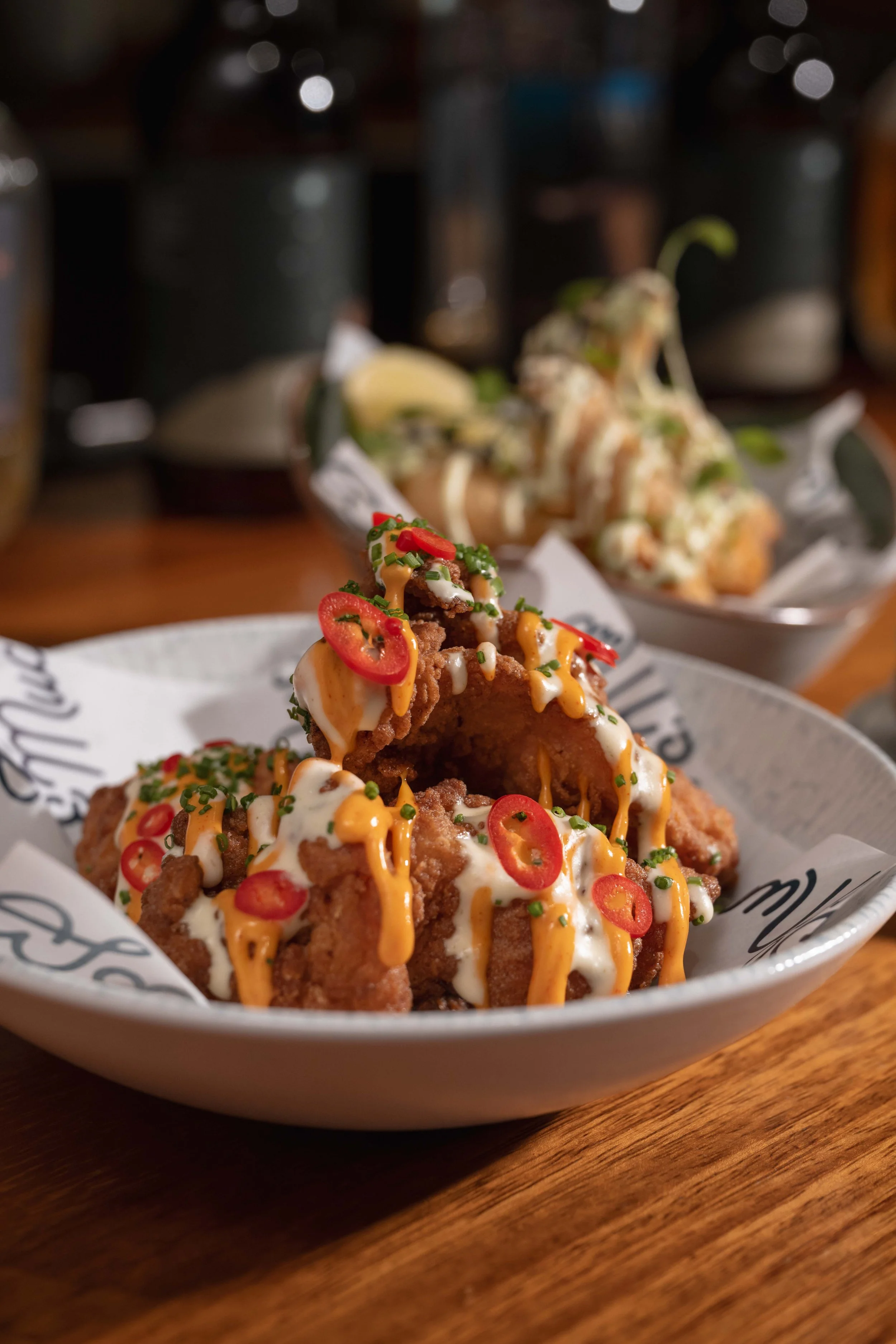 Close-up of a plate of spicy fried chicken topped with white sauce, sriracha, chopped green onions, and sliced red chili peppers, served on a paper-lined dish on a wooden table.