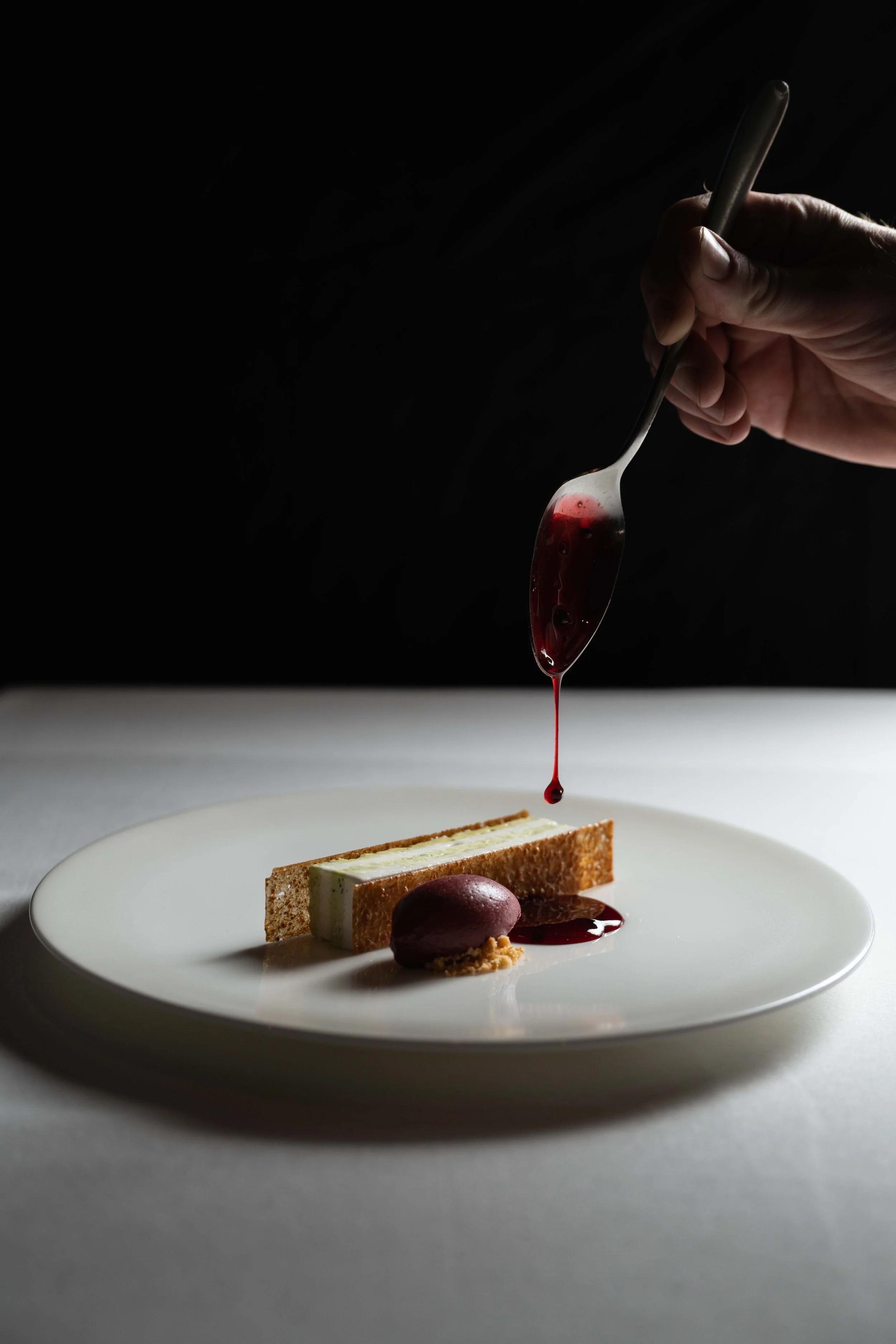 A hand drizzling red sauce over a plated dessert with a rectangular cake slice and a rounded scoop of sorbet on a white plate against a dark background.