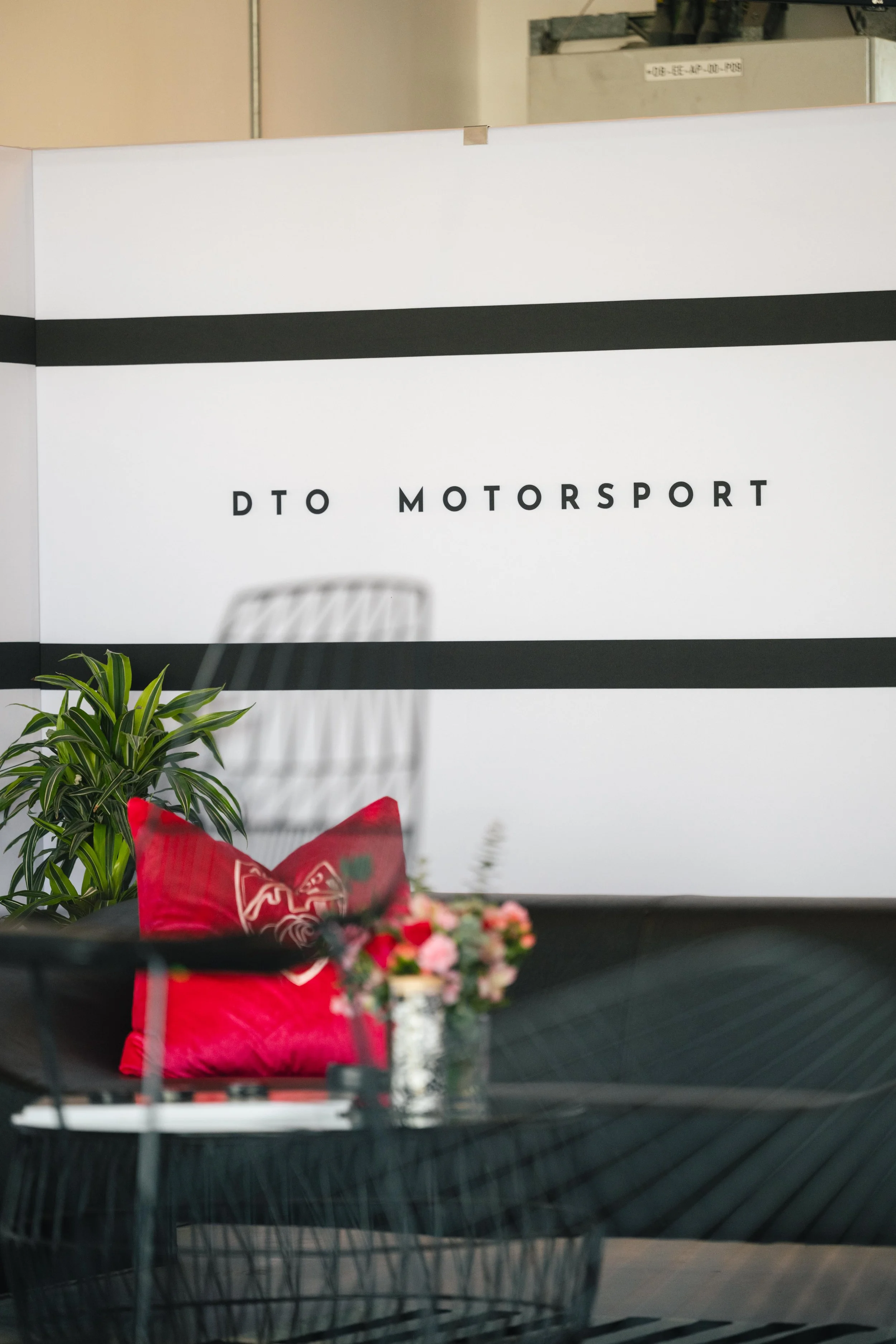 Indoor scene with a large white sign that reads 'DTO MOTORSPORT', a black wire chair with a red pillow, a small table with a vase of pink flowers, and a green potted plant.