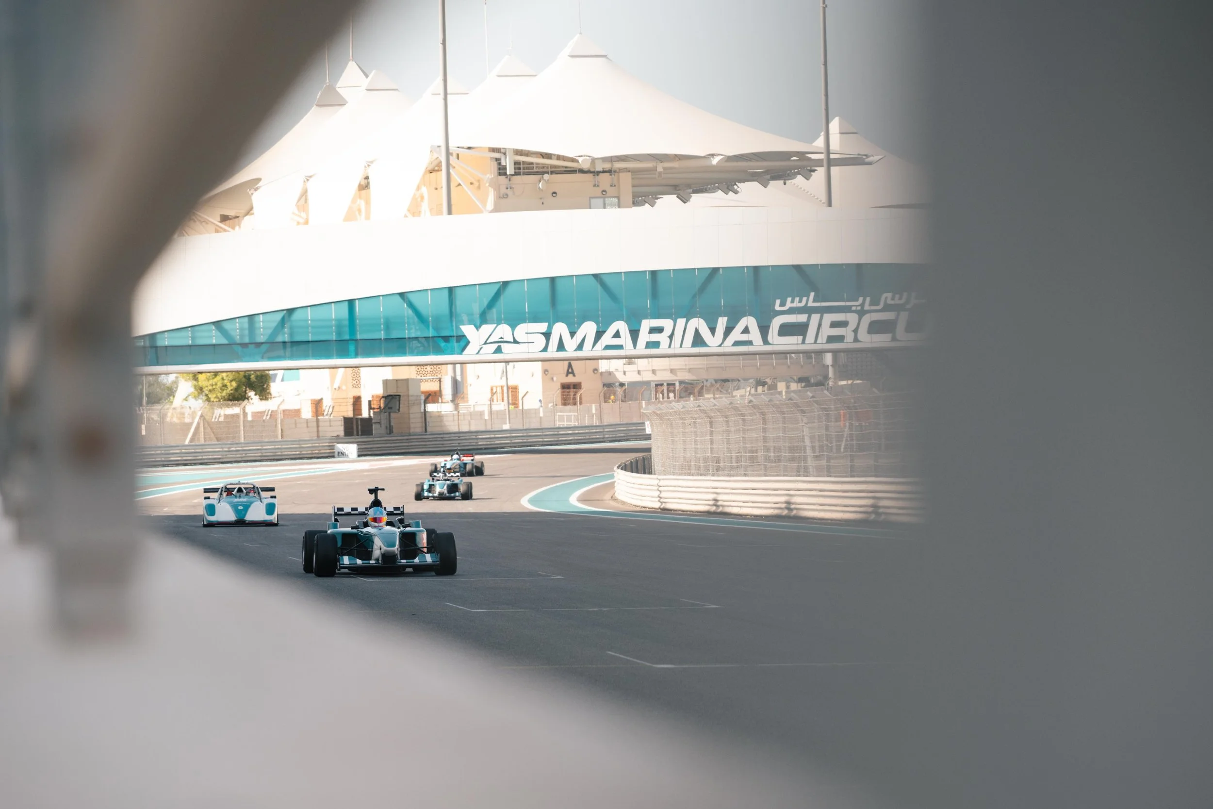 Race cars on a track at Yas Marina Circuit, with a large Yas Marina sign overhead, viewed through an opening in a barrier.