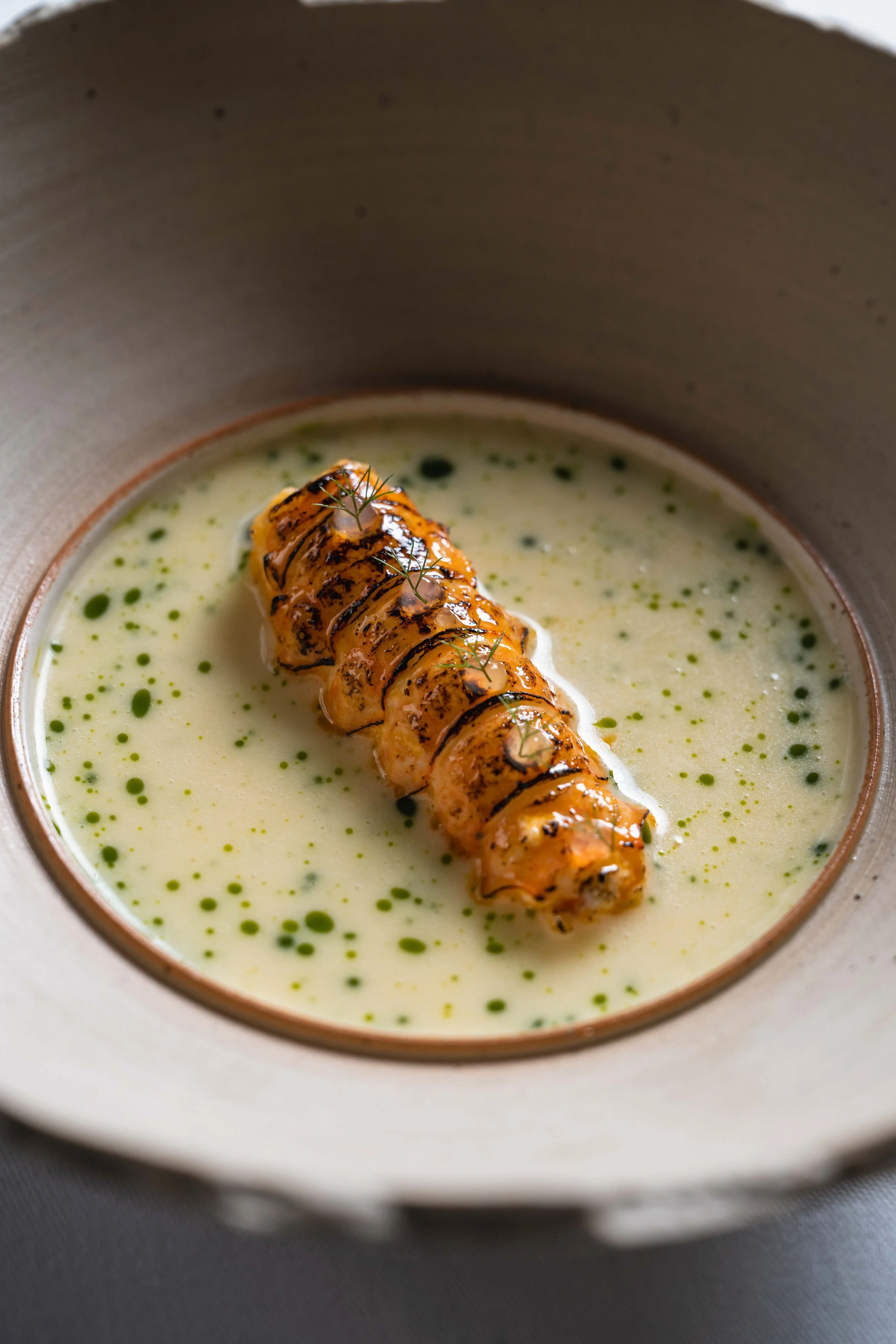 A close-up of a culinary dish featuring a grilled, glazed, and topped piece of sushi or seafood, placed in a bowl with a creamy, green sauce garnished with herbs and oil droplets.