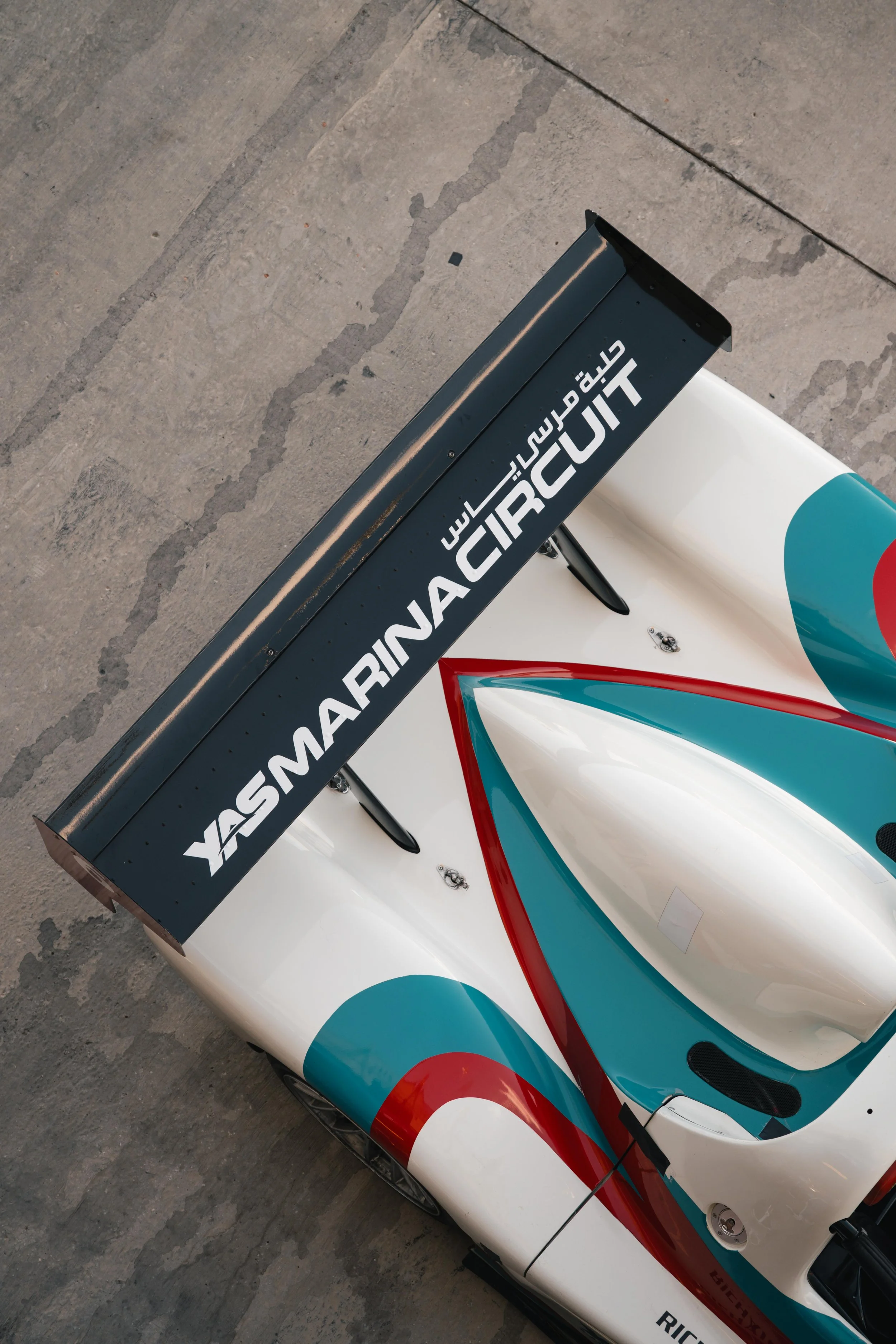 Top view of a race car with a large rear wing displaying 'YASMARINAS MARINE CIRCUS' on it, featuring a colorful turquoise, red, and white design.