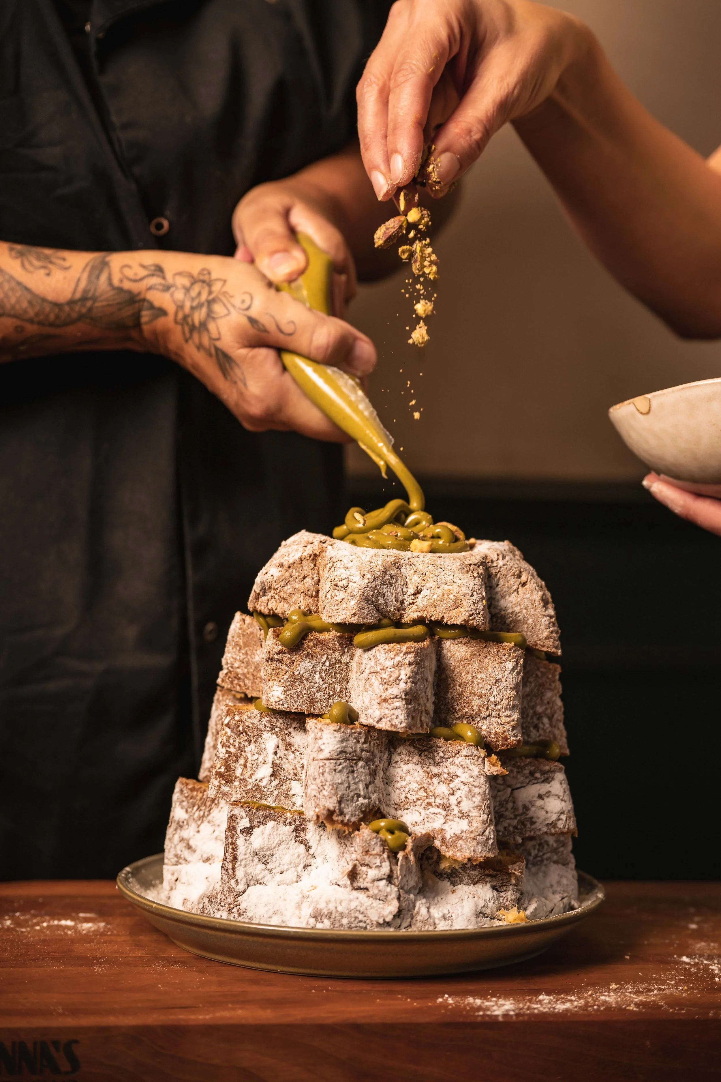 A person with tattooed arms is pouring green frosting or sauce over a stacked, cross-cut loaf of bread covered in flour, on a wooden surface.