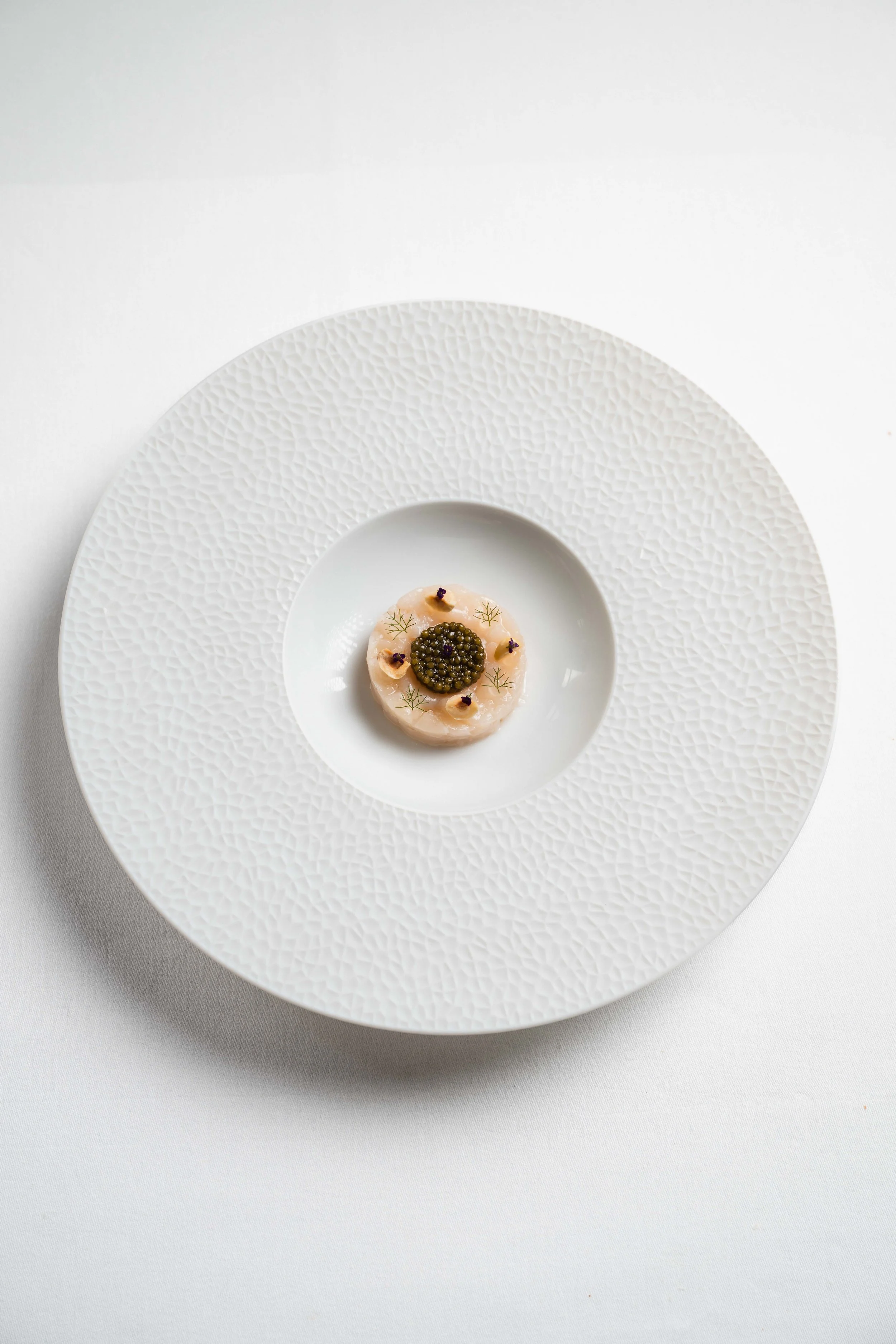 A gourmet dish presented on a large textured white plate, featuring a small, round portion topped with caviar and garnished with edible flowers.