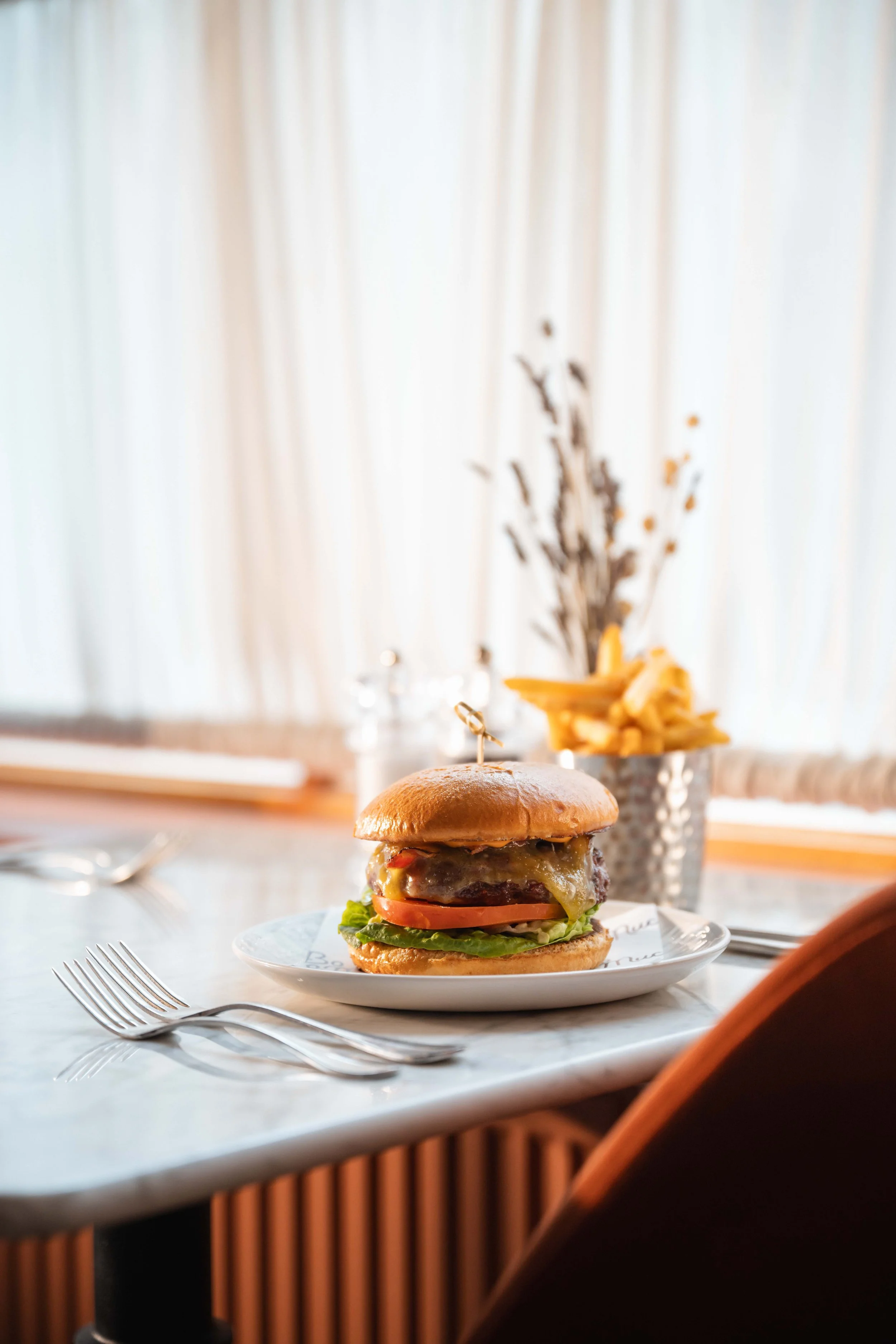 A cheeseburger with lettuce, tomato, melted cheese, and a beef patty on a bun, served on a white plate with two forks beside it. In the background, there is a small container with French fries and a blurred floral display.