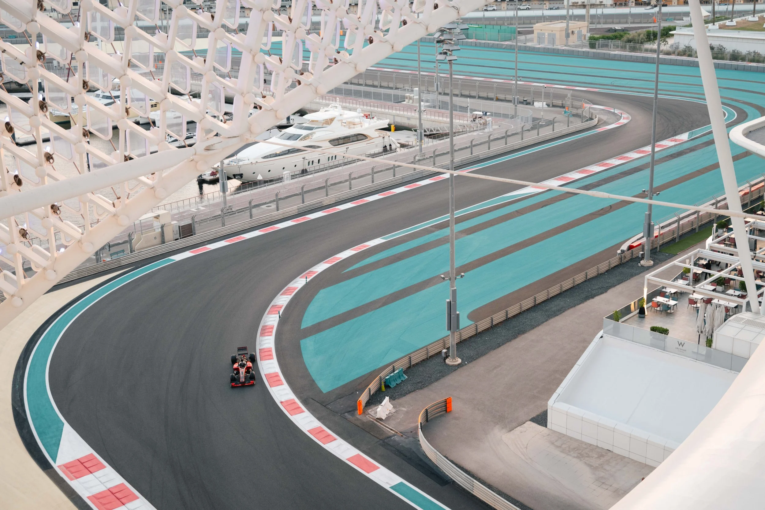 A race car driving on a race track with blue painted sections, curving on a race circuit with barriers and infrastructure surrounding it.