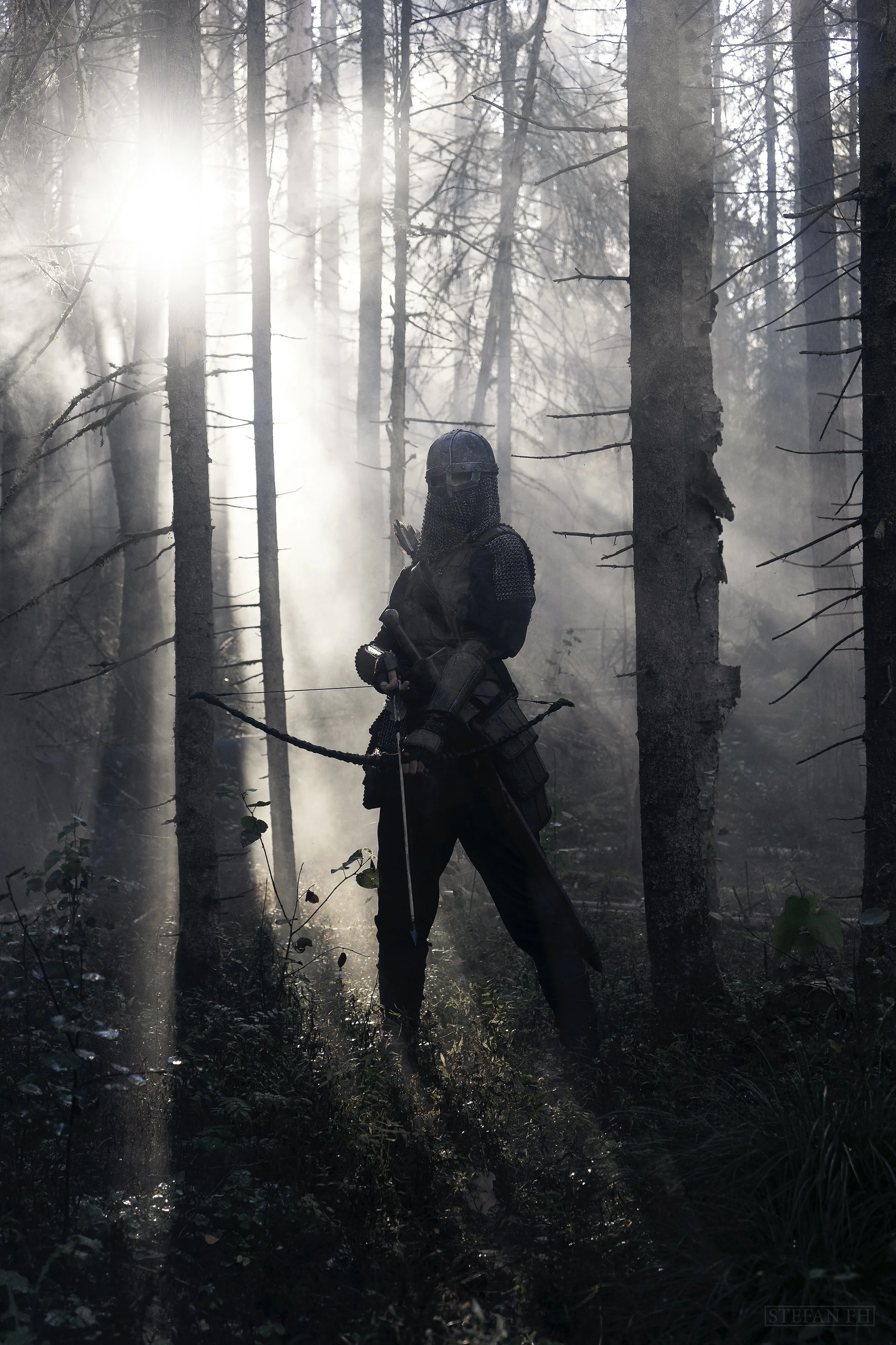 A person dressed in medieval knight armor, holding a sword, standing in a foggy forest with tall trees and sunlight shining through.