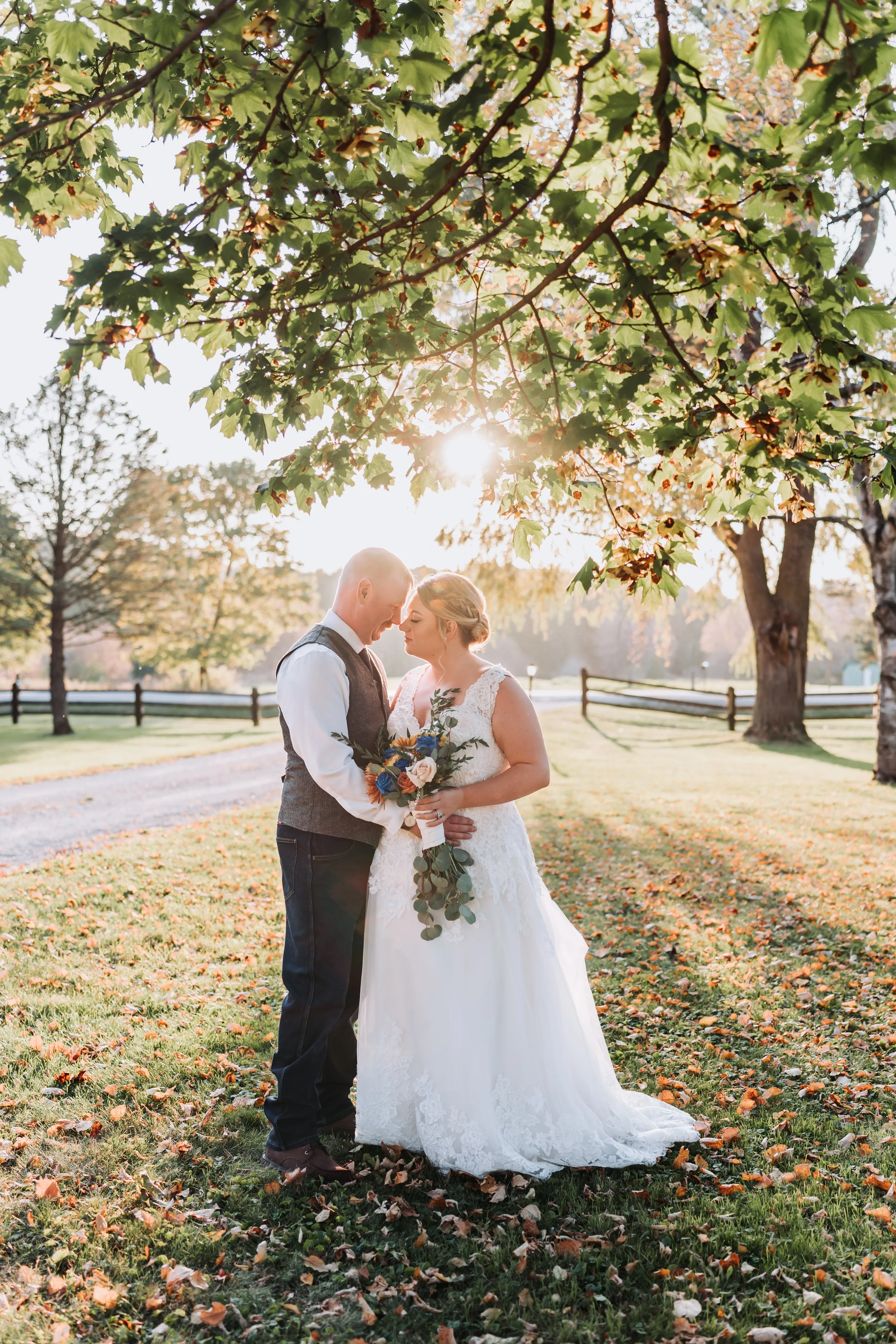 Documentary-style wedding photography of a bride and groom standing under a tree at sunset surrounded by autumn leaves at On The Ridge Events by Maple & Clover.