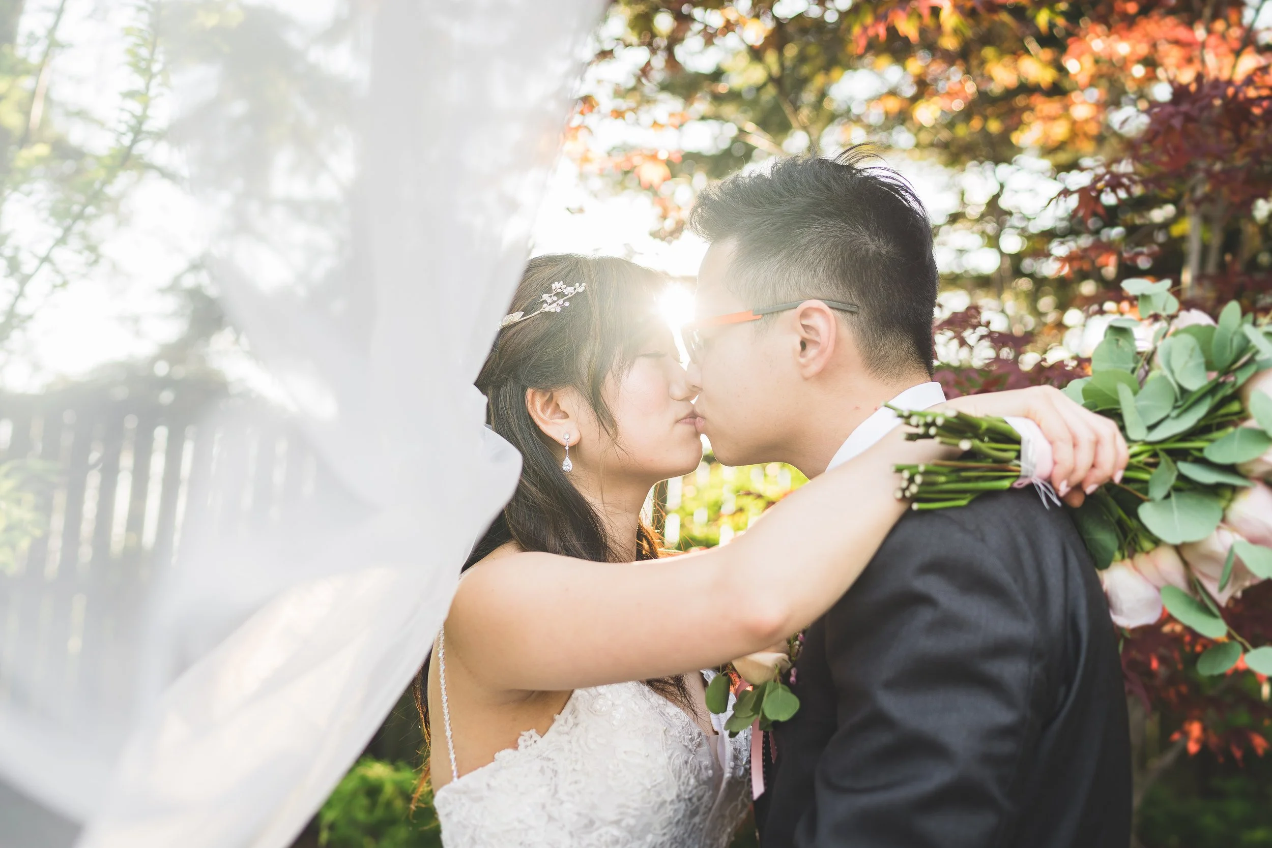 Bride and groom kissing outdoors with sunlight shining behind them, surrounded by greenery and autumn leaves, bride holding a bouquet of flowers.