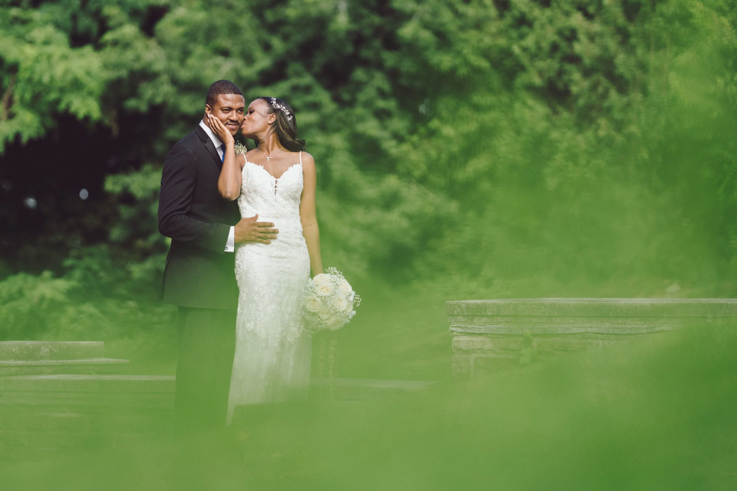 Documentary-style wedding photography of a bride kissing the groom on the cheek outdoors with green trees in the background at the Glen Abbey Golf club, by Maple & Clover.