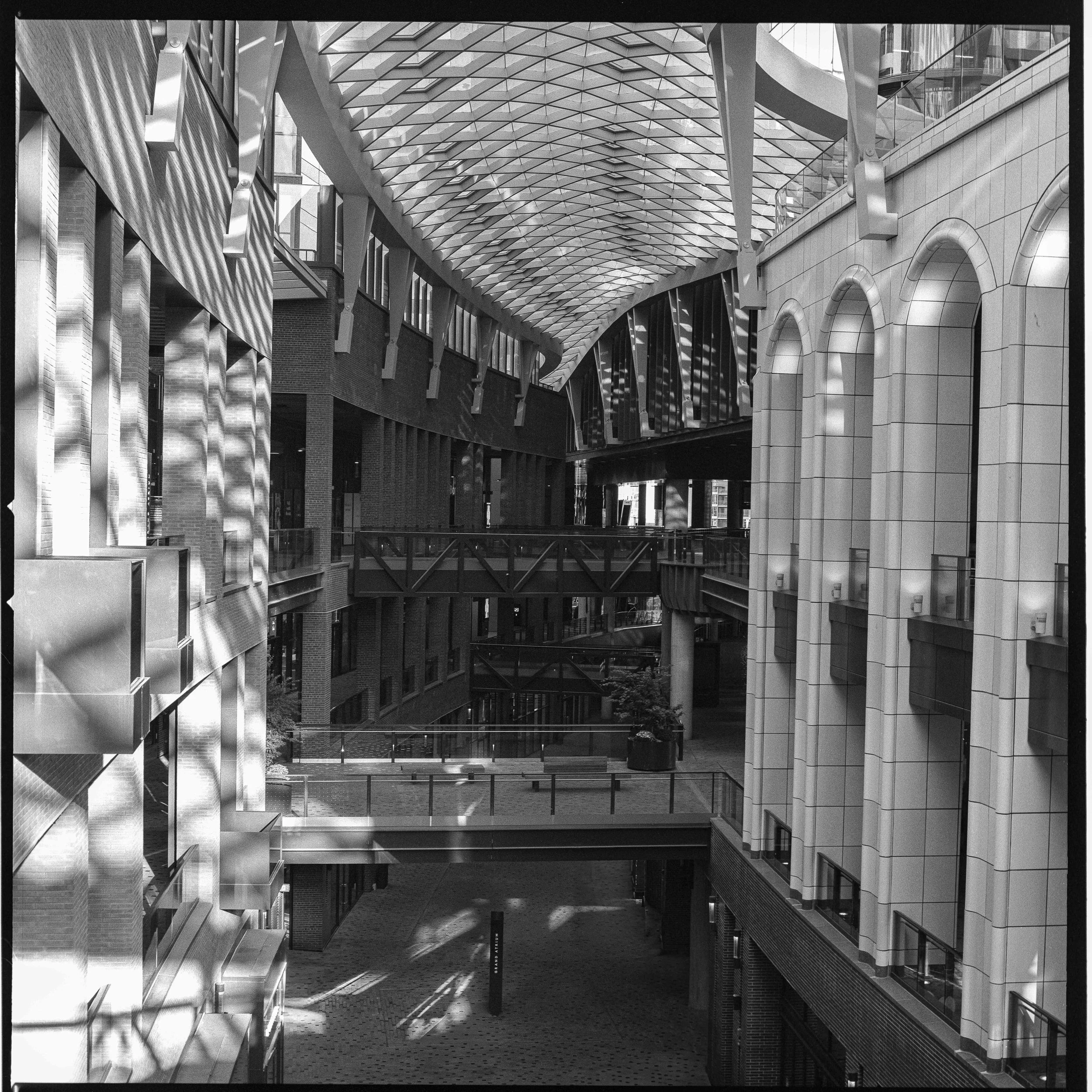 Interior view of a modern multi-level shopping mall with sunlight creating patterns, captured in a street photography style by David Stokes.