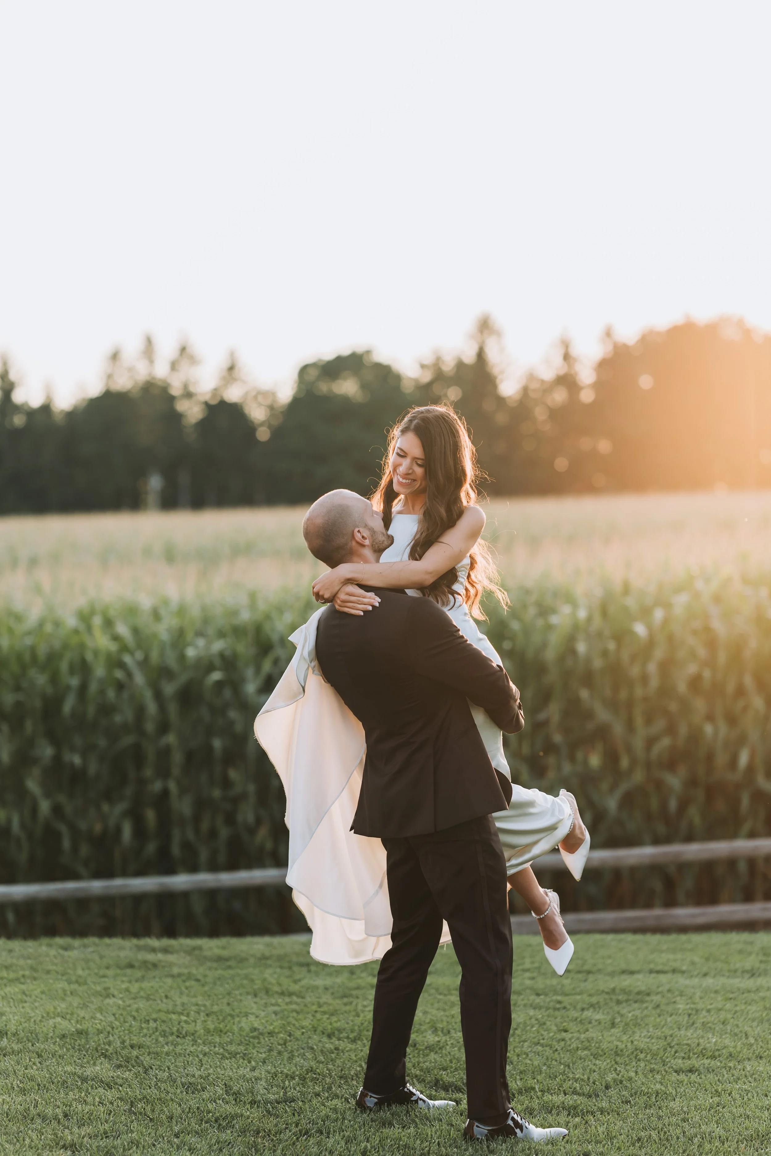 Documentary wedding photography of a couple in Barrie by Maple & Clover couple in formal attire, with the man lifting the woman, smiling at each other in a field at sunset.