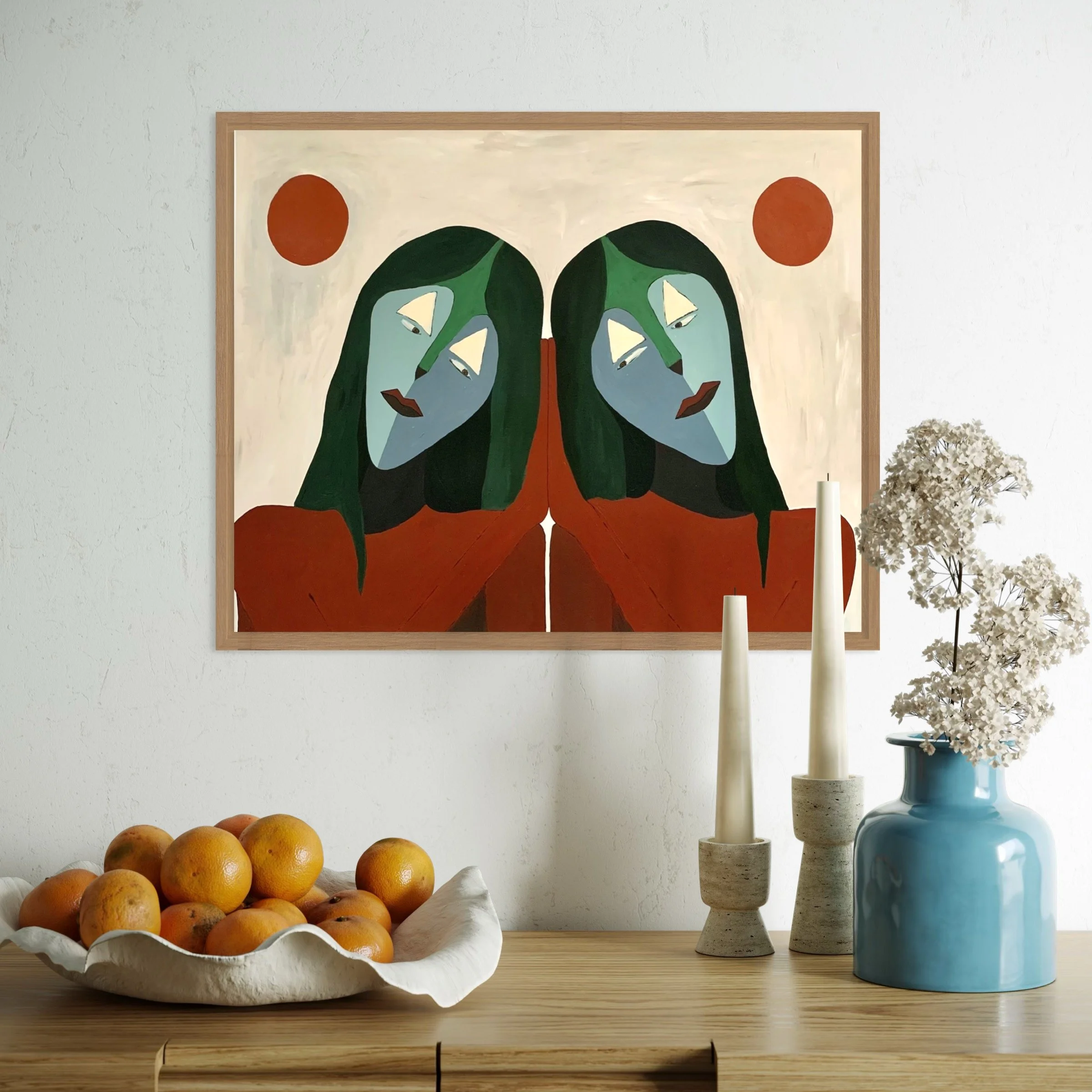 A table with a ceramic bowl of oranges, a glass of vanilla ice cream, a blue glass vase, and a white cylindrical candle holder with candles. Behind the table is a white wall with a framed abstract artwork featuring green humanoid figures and red circ