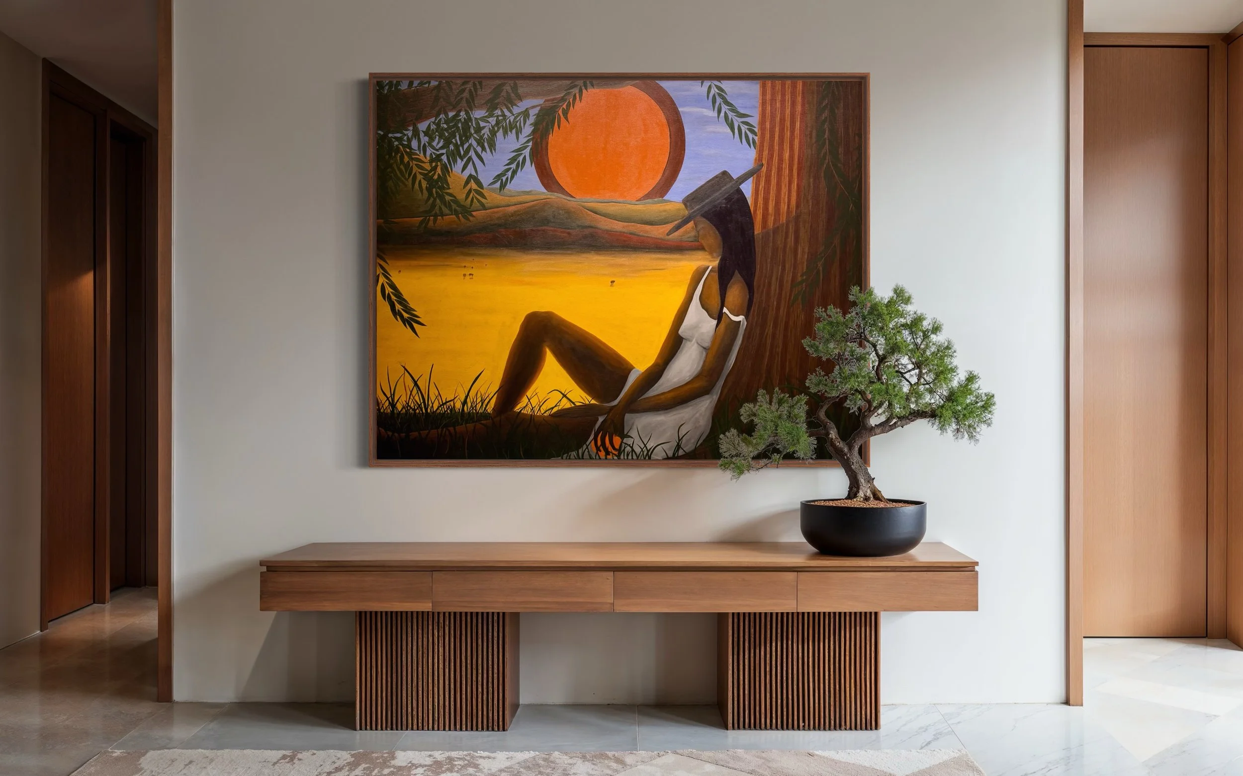 A painting of a woman sitting under a tree with a large sun in the background, in warm tones, mounted on a white wall above a wooden console table with a bonsai tree in a black pot on top.