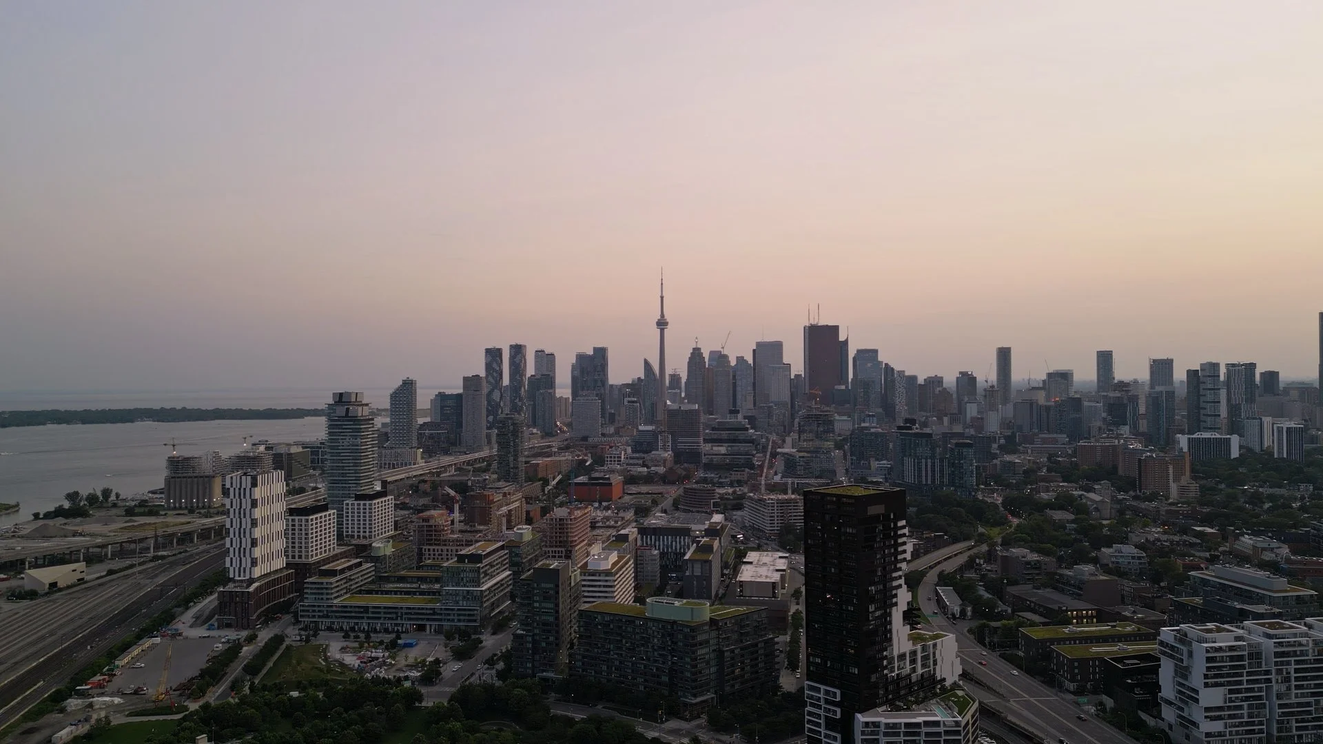 A city skyline at dusk featuring numerous tall buildings, with the prominent CN Tower in the center, and a river to the left. The sky is light with a pastel hue.