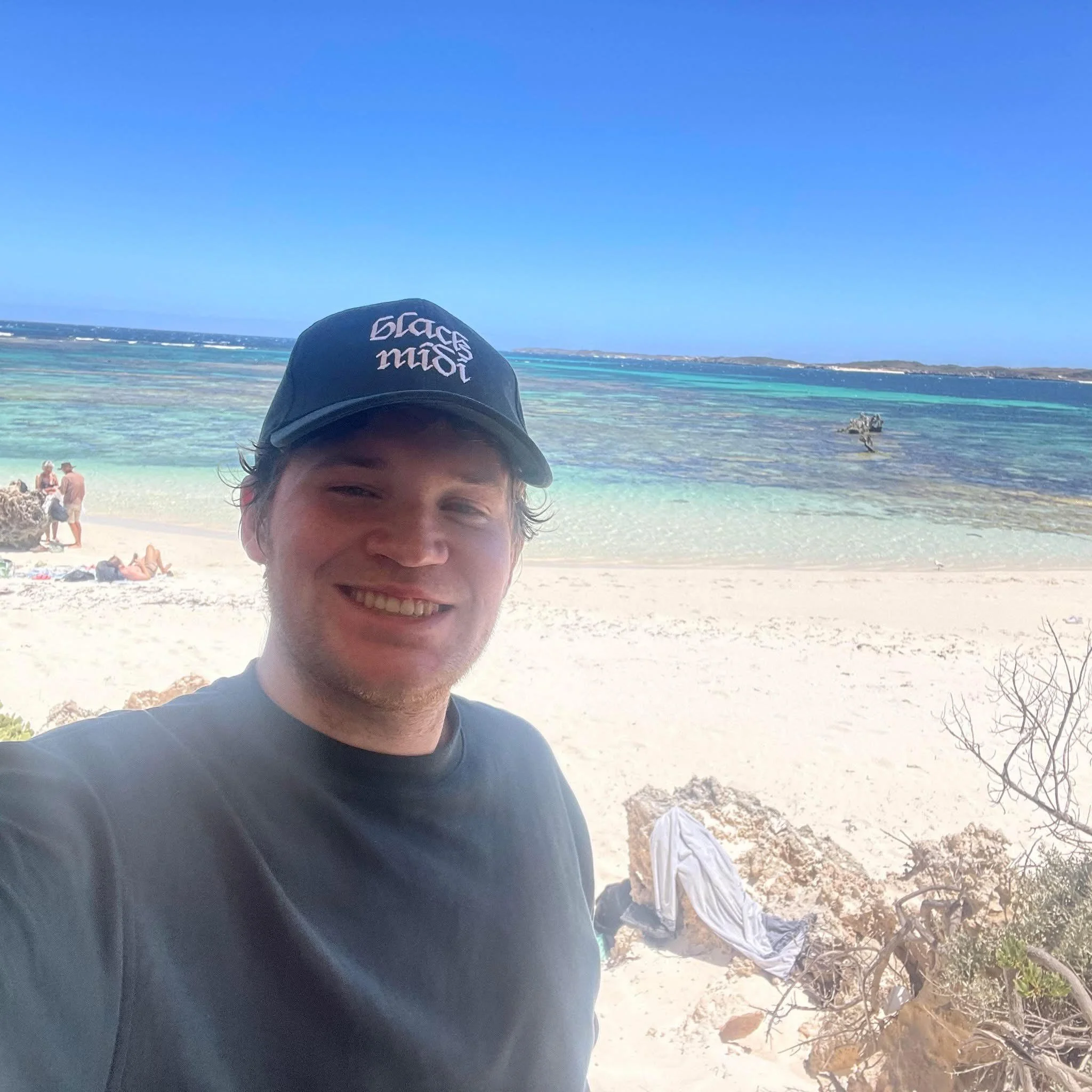 A photo of Alec smiling to the camera. He is on a beach with sand and water in the background, and is wearing a tshirt and cap.