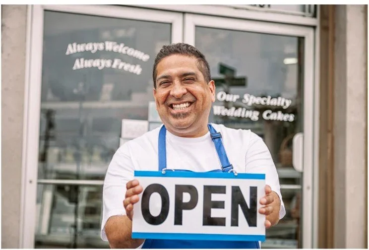 A smiling man, holding a sign that says, "OPEN" in his hands, in front of his wedding cake store