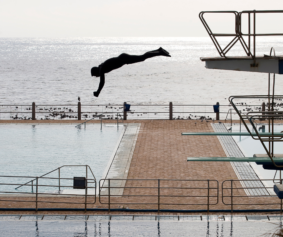 A man diving off of a high dive, into a pool, outdoors.