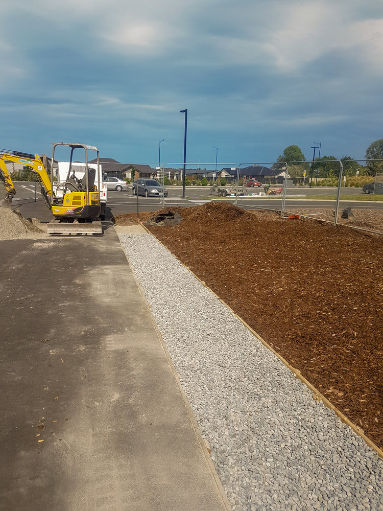  A concrete area recently installed alongside a perfectly edged gravel path with wood edges. Alongside a fresh bark pile being prepared for plants as a garden area. A Hannon Civil Digger rests against a pile of gravel with Christchurch, Merivale hous