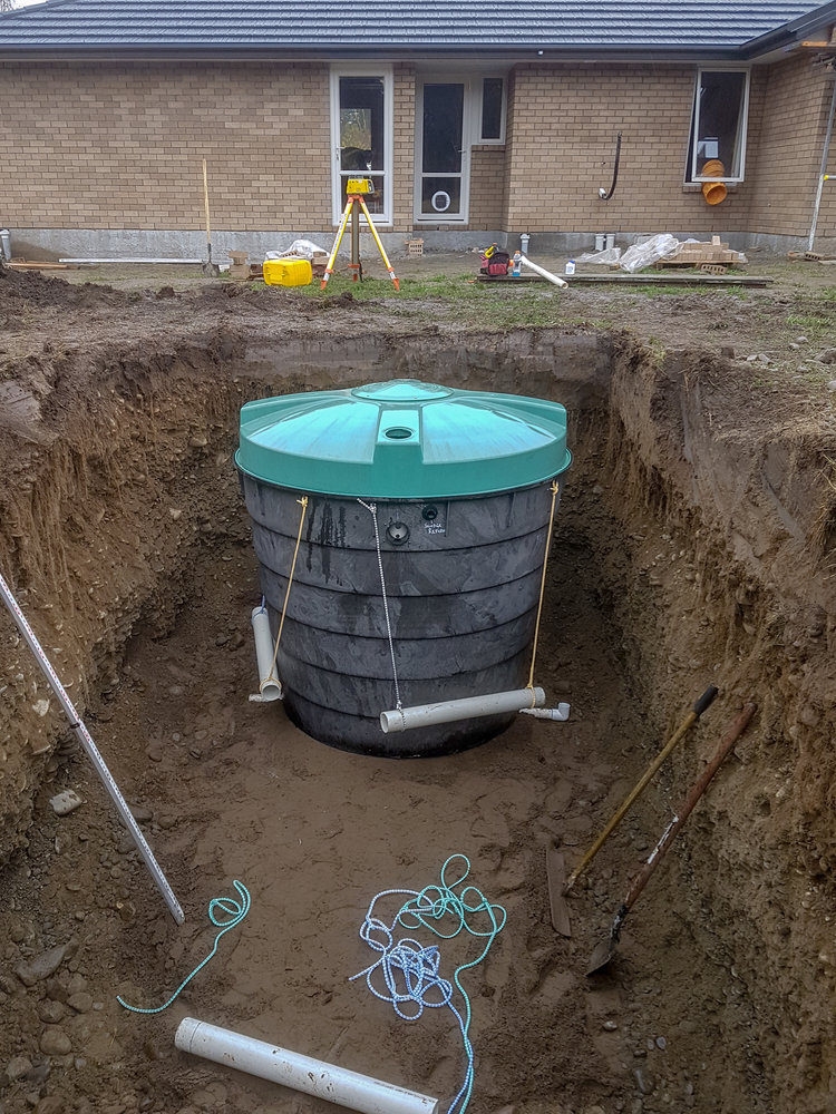  a Septic tank sits in a ditch ready to be installed with a Specra precision laser measuring the ground. both drainlaying and civil construction tools are in the ditch and in the background including shovels, pipes, wheelbarrows 