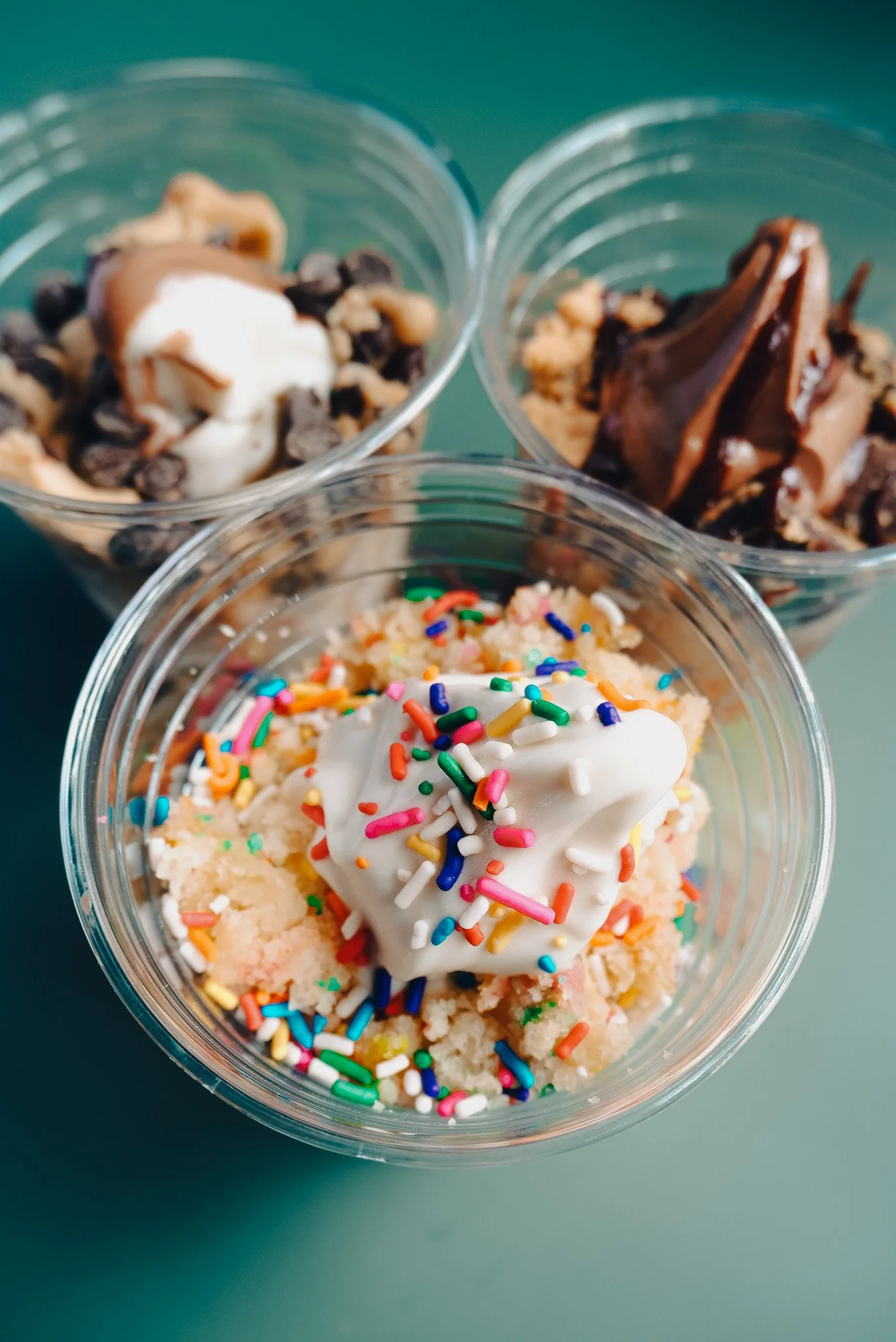  Three clear cups filled with soft serve and toppings are placed together on a green background. The one in the front has funfetti cake as a topping and rainbow sprinkles. 