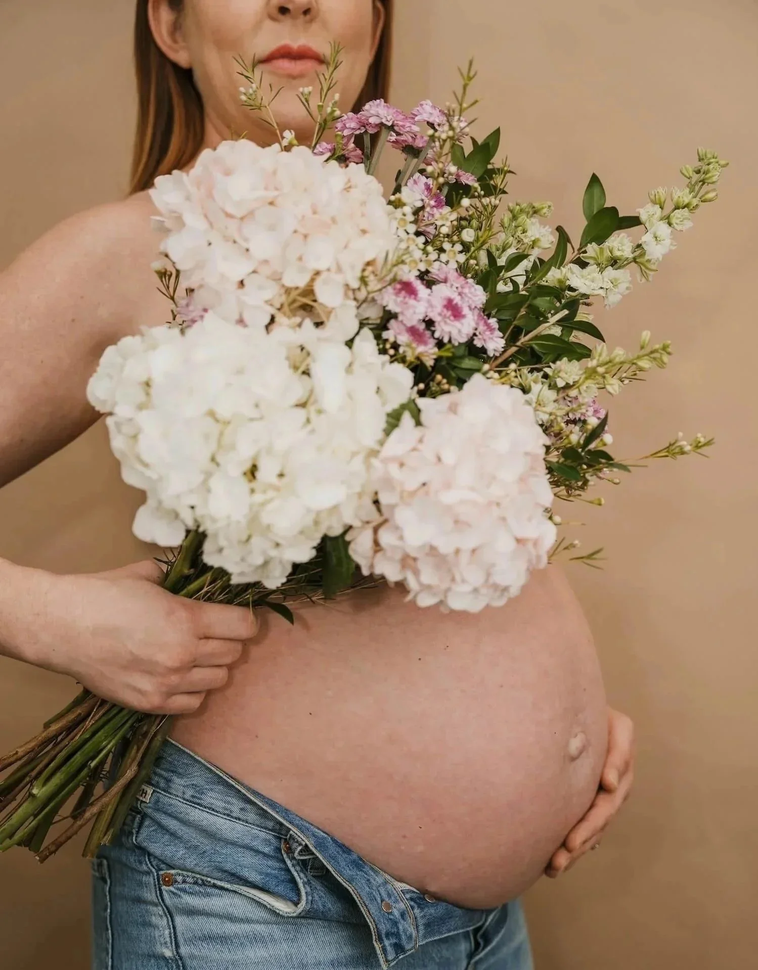 Pregnant woman standing in profile with a bear belly holding a bouquet of white and pink flowers to cover her chest. She has red hair and is wearing jeans.