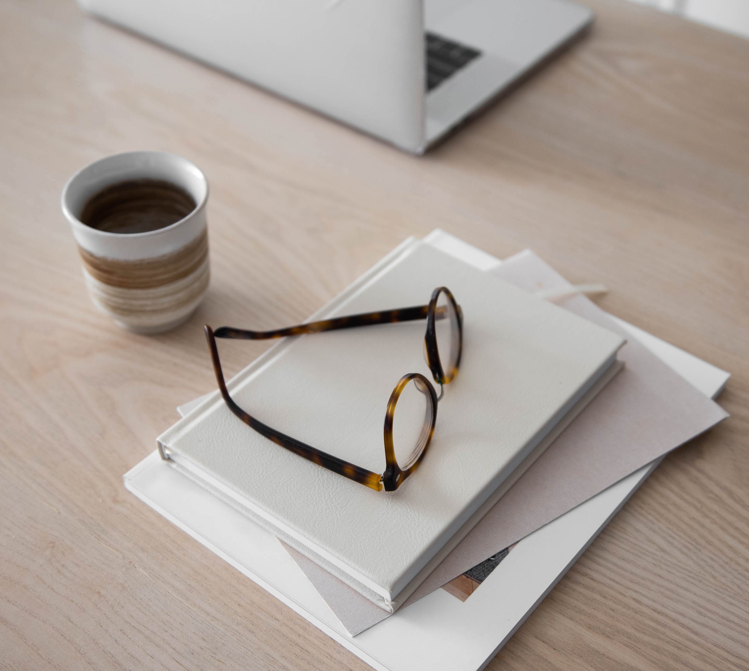 A light wood table with a stack of white books and folders with a pair of tortoise shell reading glasses atop. There's a ceramic cup of coffee and a silver laptop
