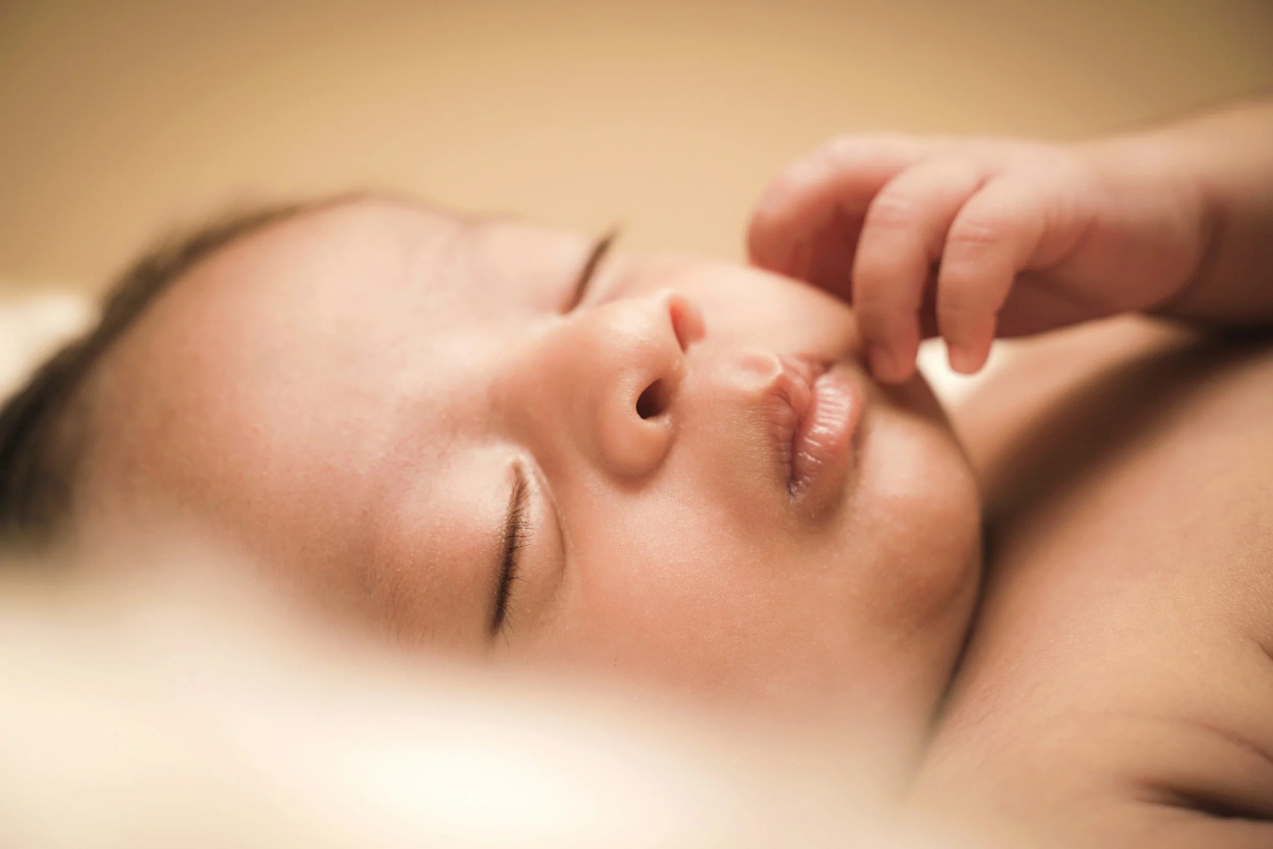 closeup of a newborn baby with eyes closed and hand up to face sleeping peacefully with a light brown background